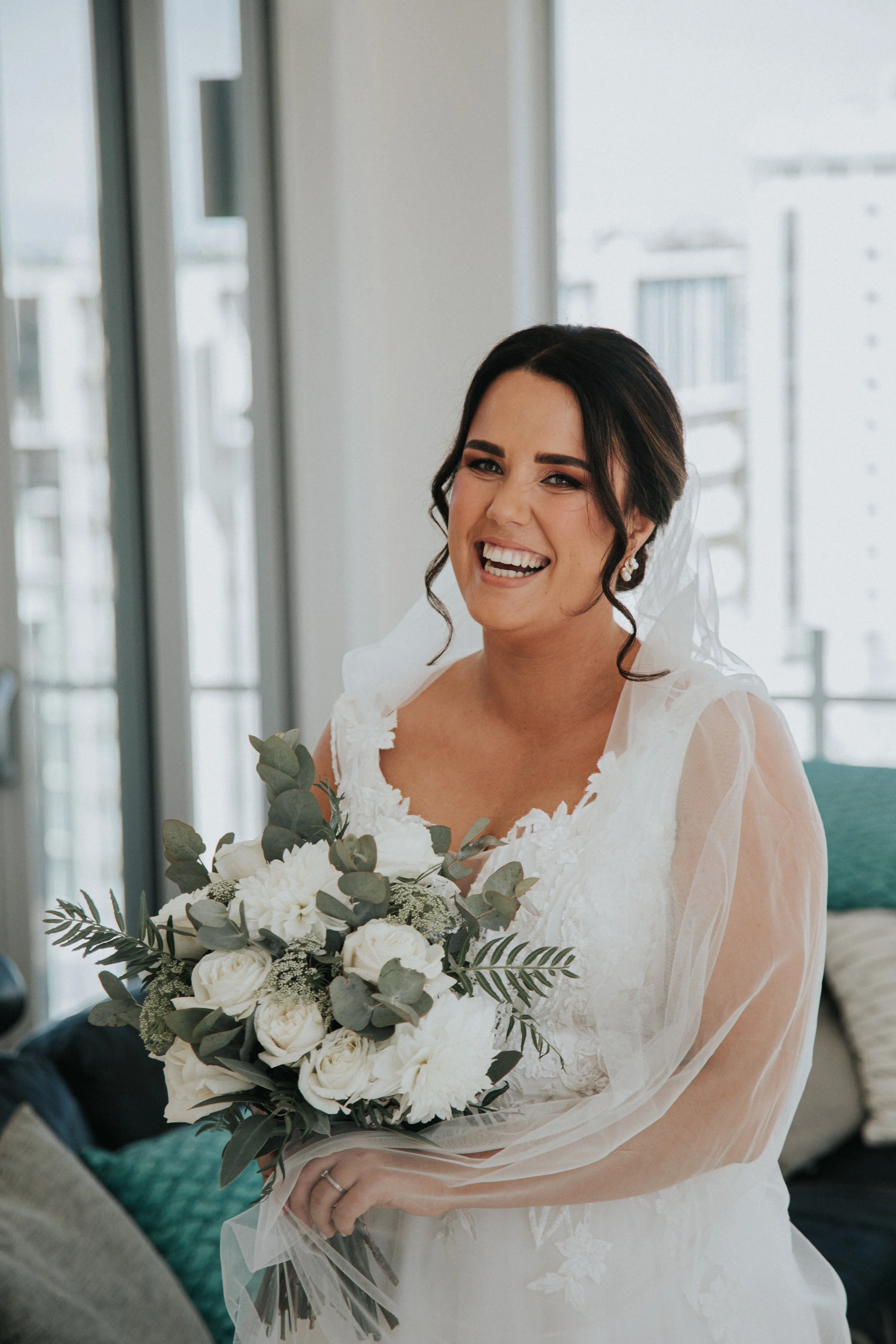A smiling bride in a white wedding dress holding a bouquet of white roses and greenery.