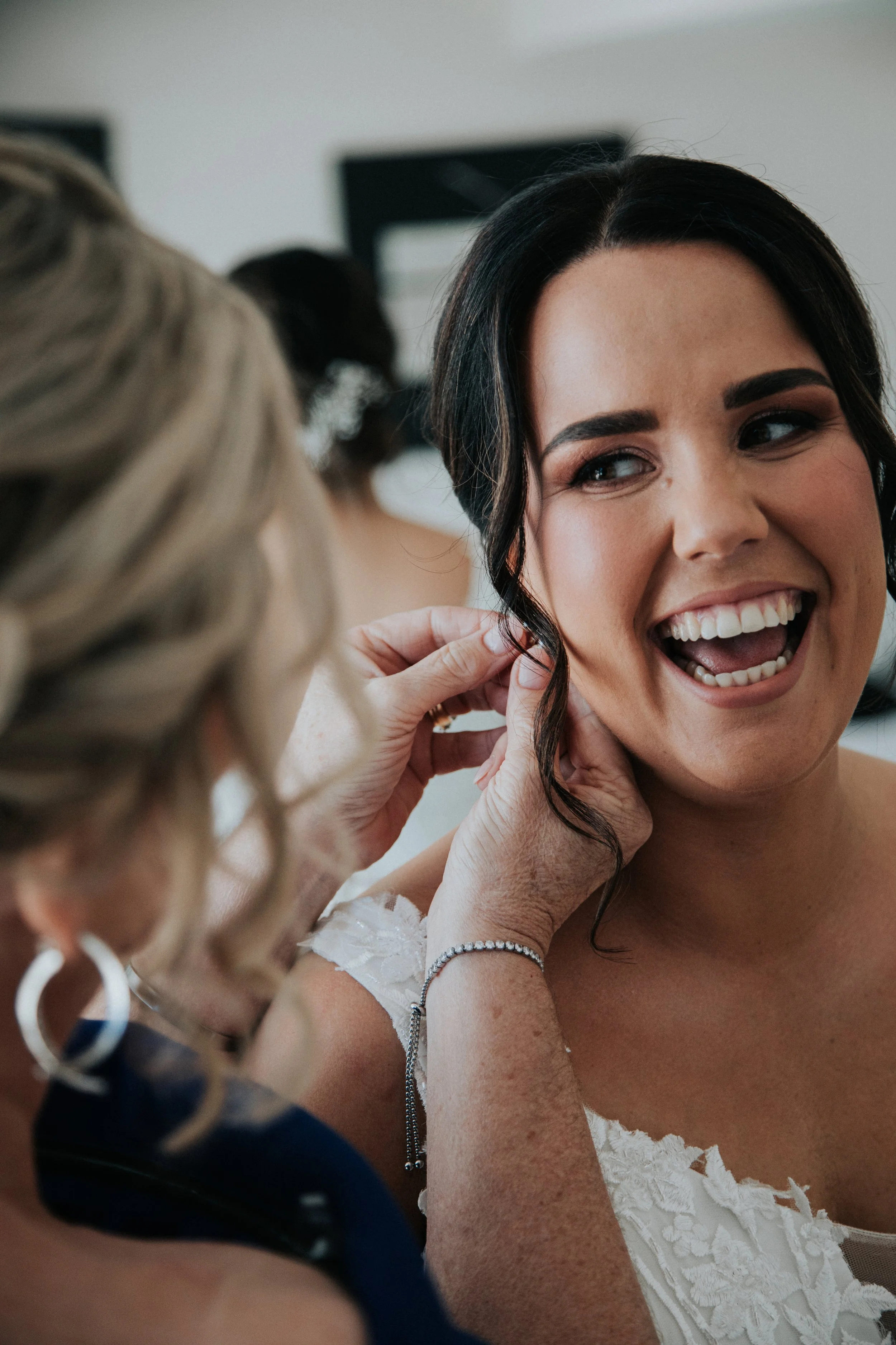 A woman with dark hair smiling as another woman adjusts her earring, with a bride's dress visible.