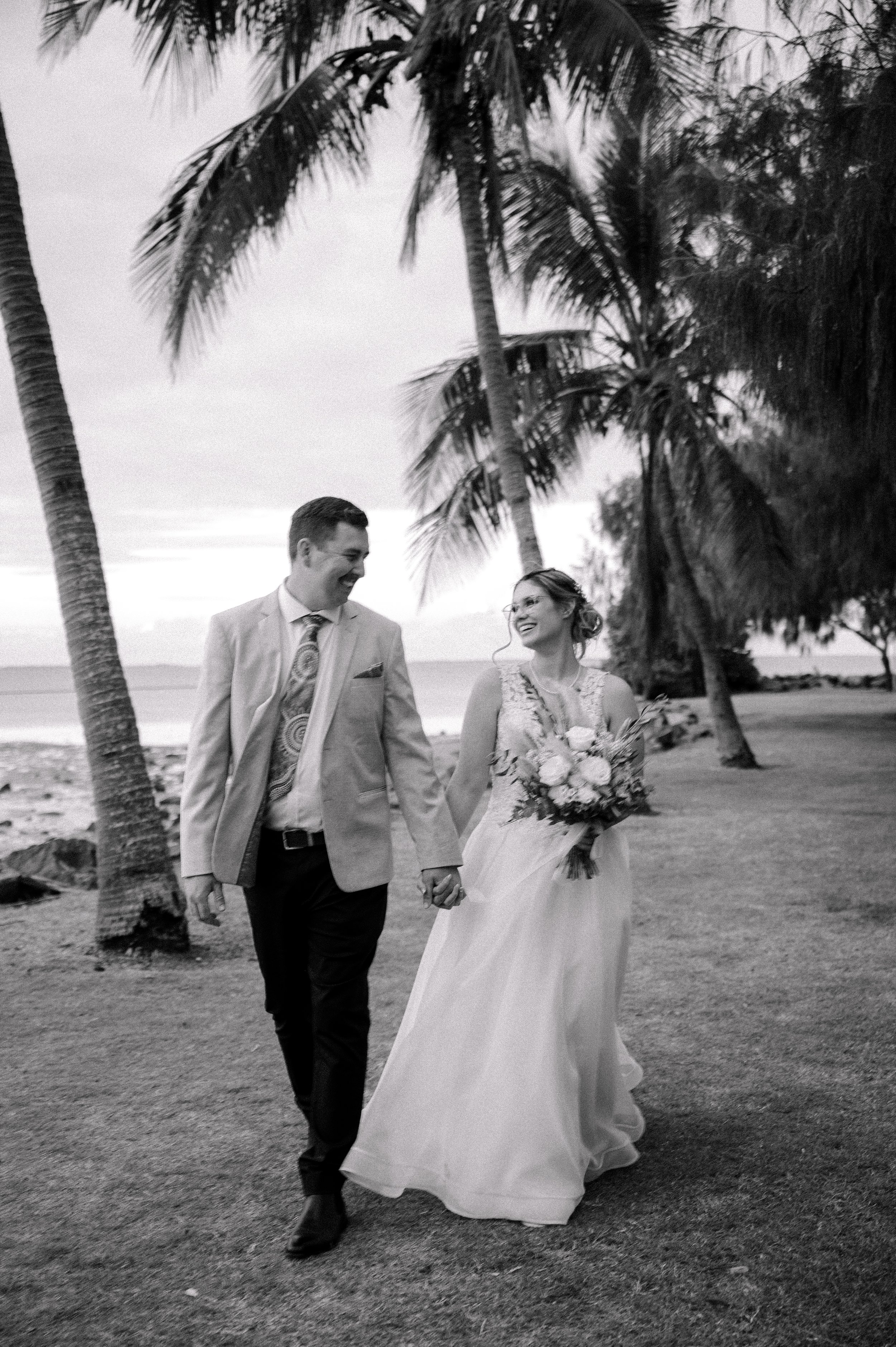 A bride and groom walking hand-in-hand on a beach, smiling at each other, with palm trees in the background, during sunset.
