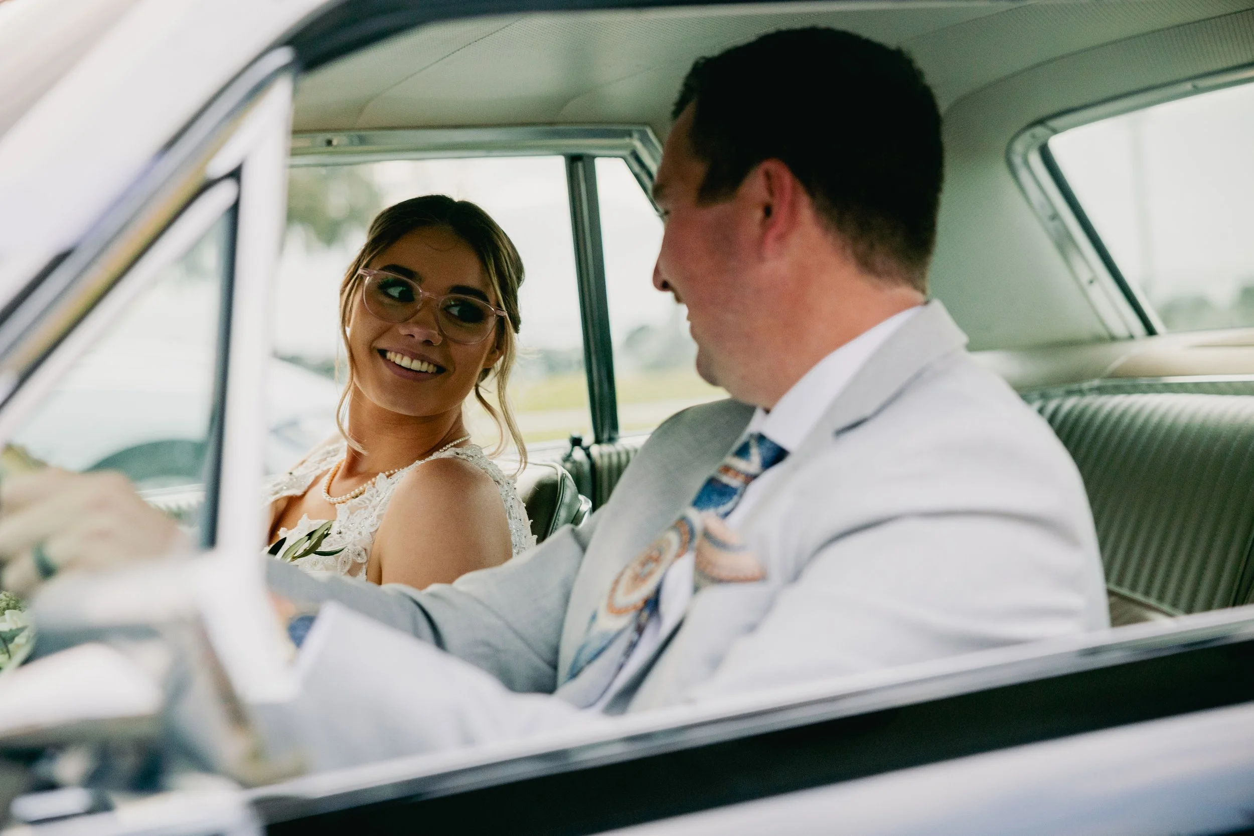 A happy couple in wedding attire sitting inside a vintage car, smiling at each other.
