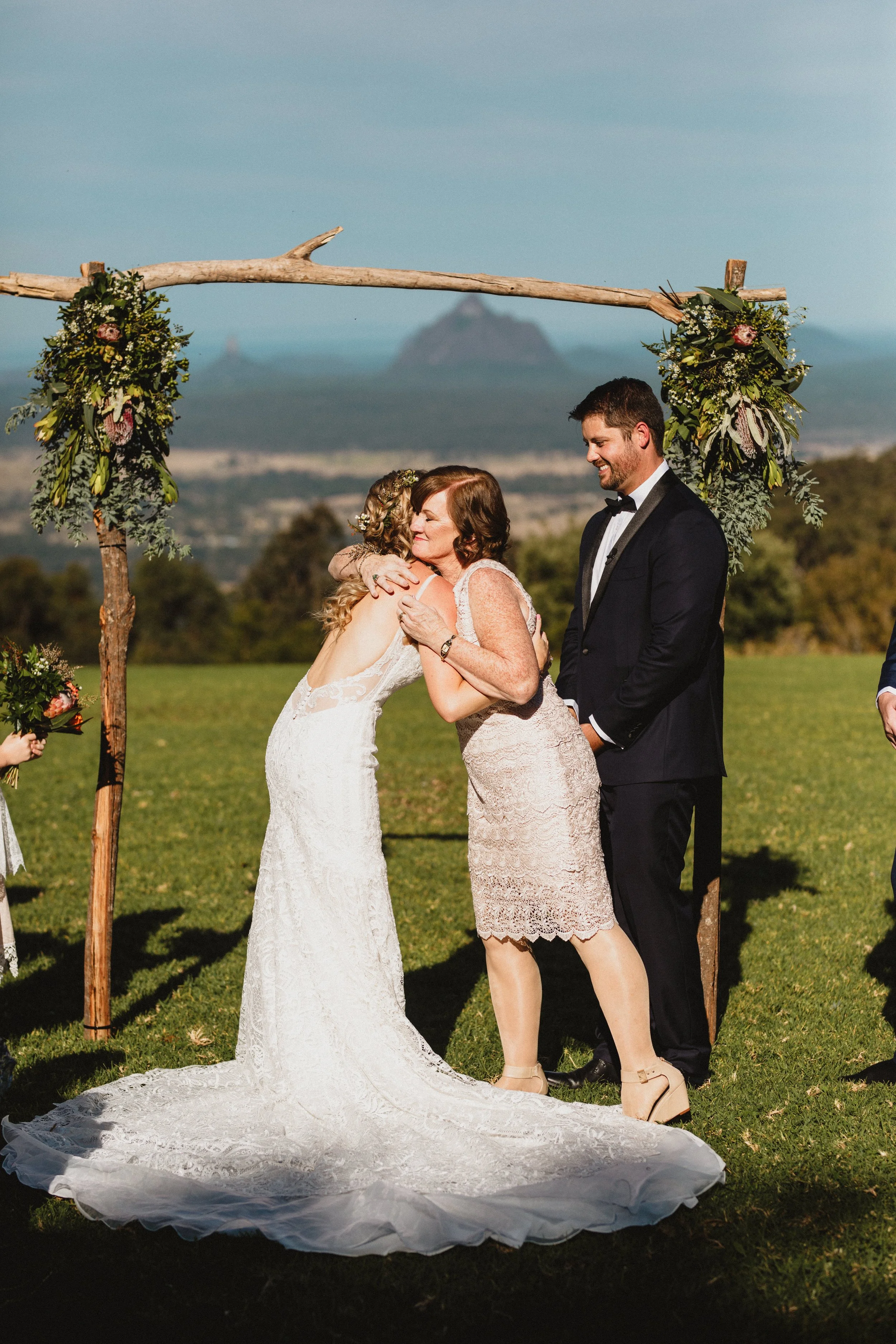 A bride and her mother hugging at an outdoor wedding ceremony under a wooden arch adorned with greenery and flowers, with a mountain and landscape in the background.