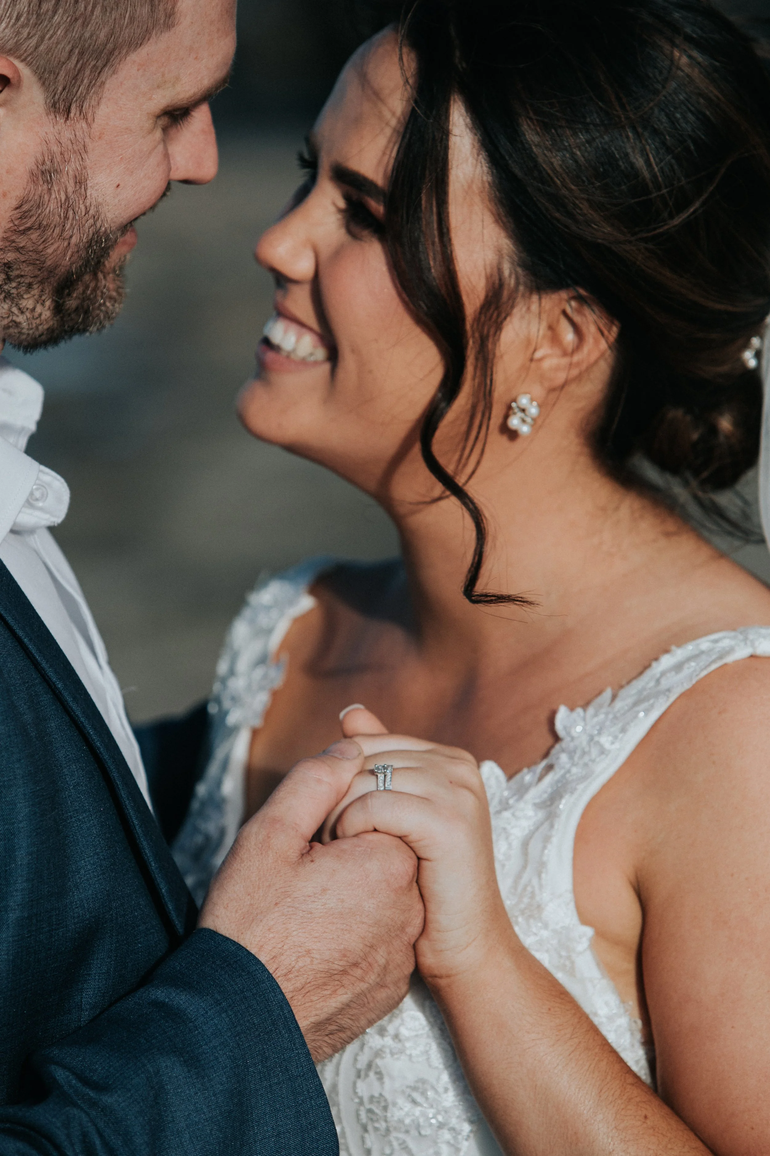 A bride and groom holding hands and smiling close to each other, dressed in wedding attire, outdoors during the daytime.