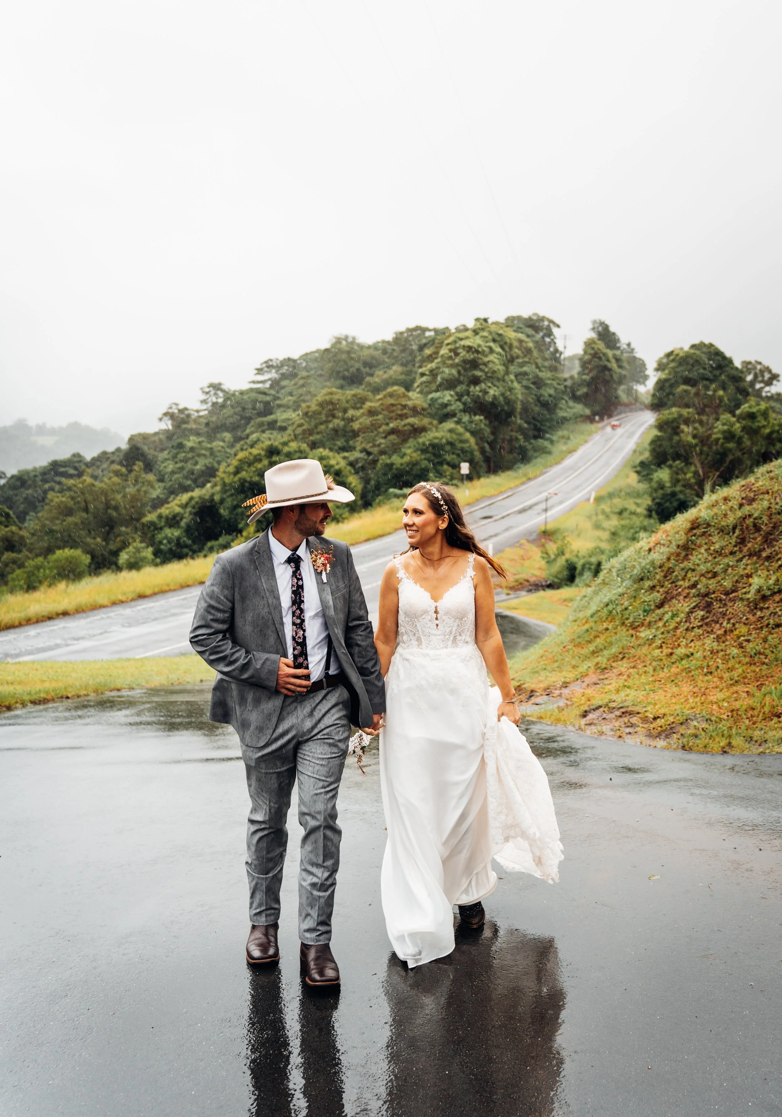 A newlywed couple walking hand in hand on a wet road on a rainy day, surrounded by lush green hills and trees.