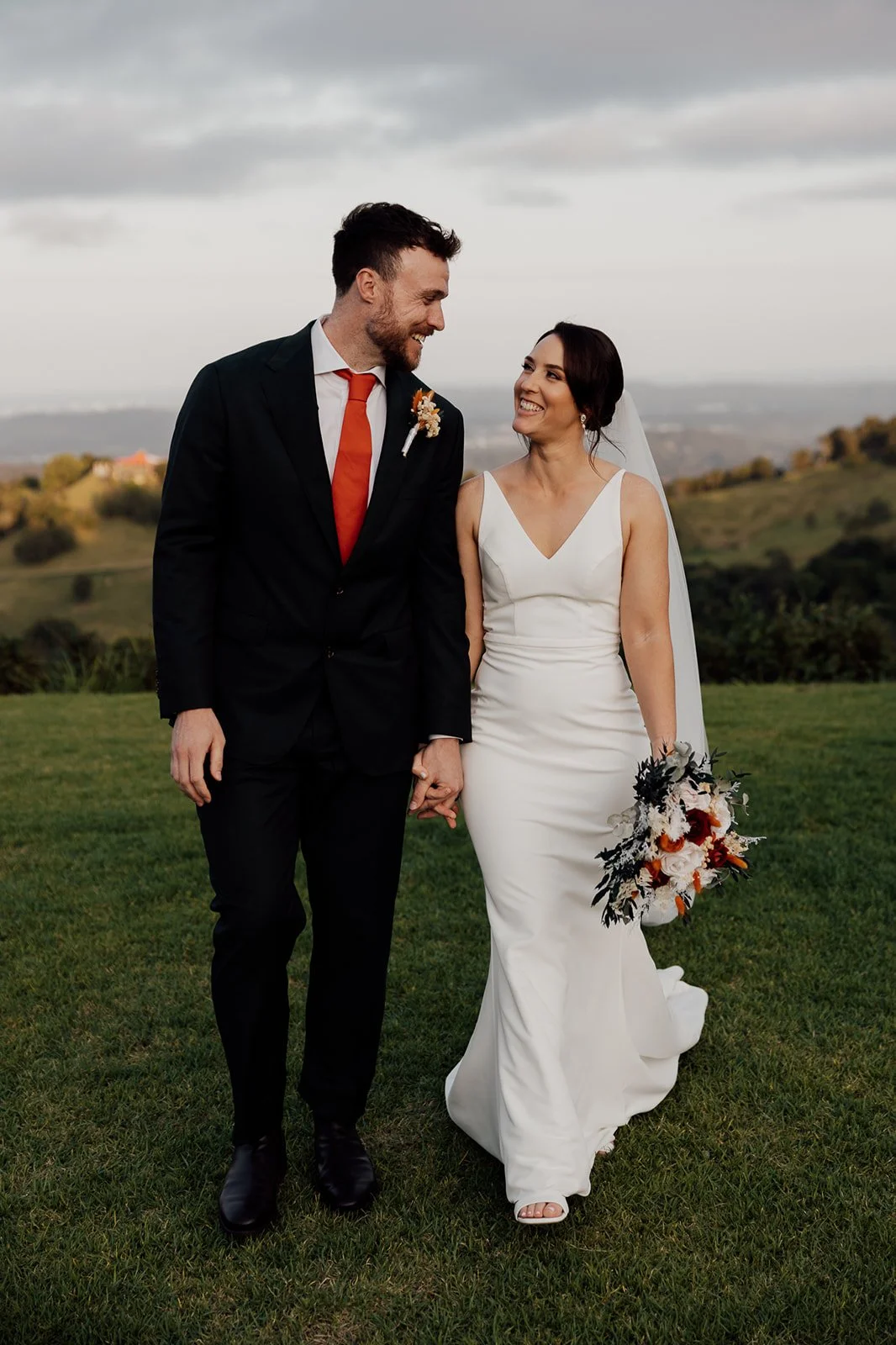 A bride and groom walking hand in hand on a grassy field, smiling at each other. The groom is dressed in a black suit with a white shirt and a red tie. The bride is wearing a white sleeveless wedding gown, holding a bouquet of flowers, and wearing a 