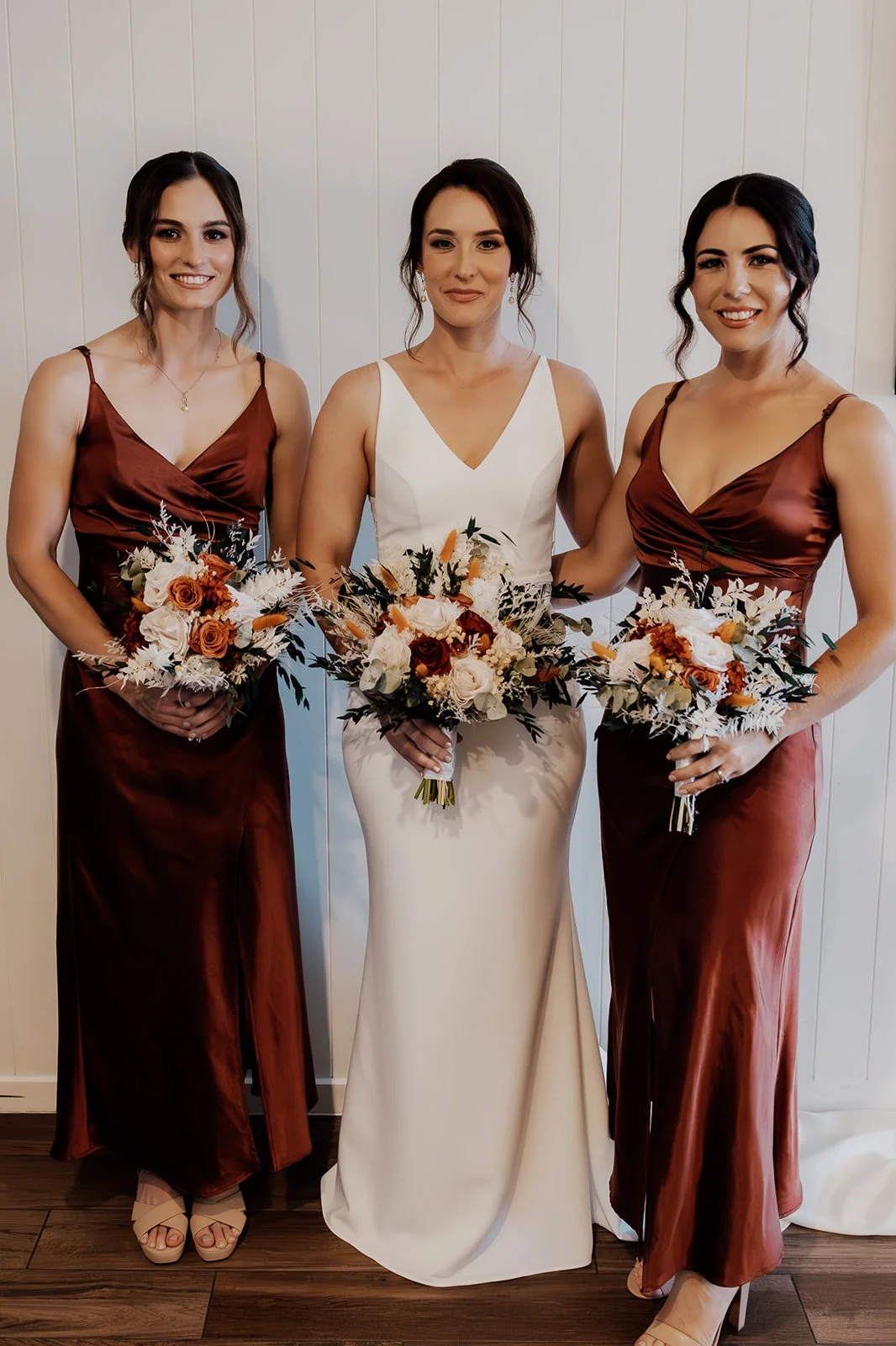 Three women dressed in formal attire, holding bouquets of flowers, standing against a white paneled wall.