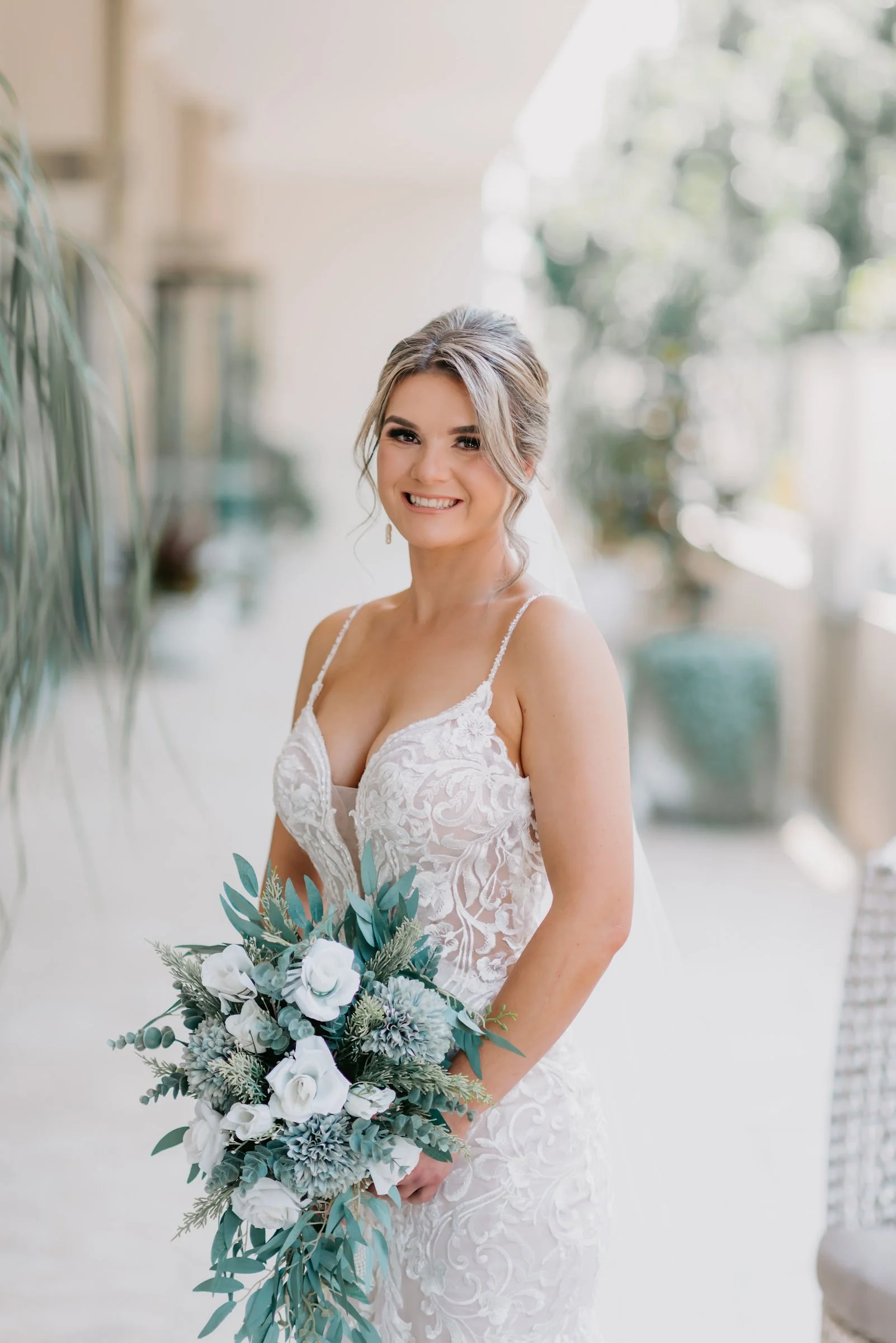A bride in a lace wedding dress holding a bouquet of white and blue flowers, standing indoors with a blurred background.