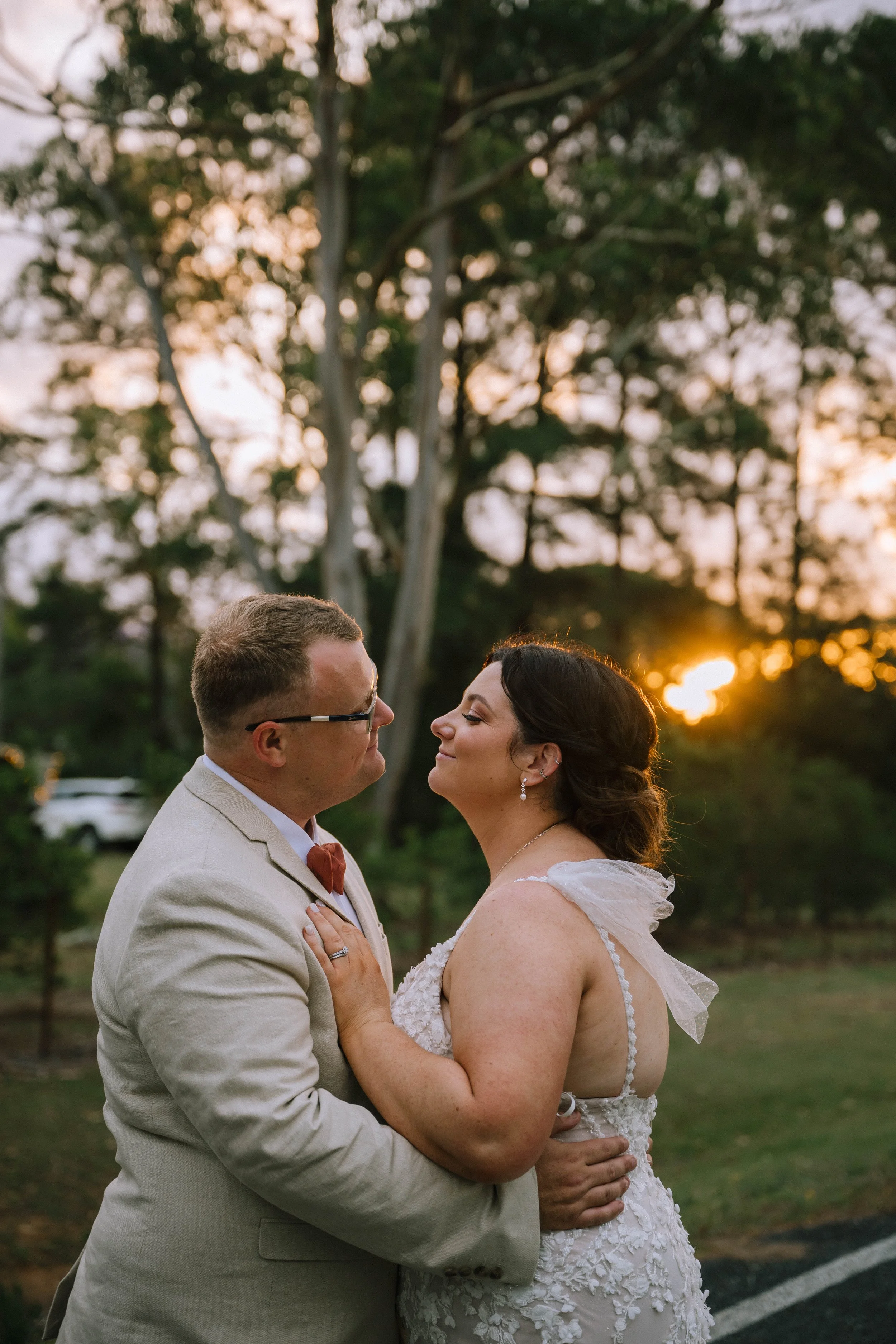A bride and groom share a romantic moment outdoors during sunset, with trees and a park in the background.
