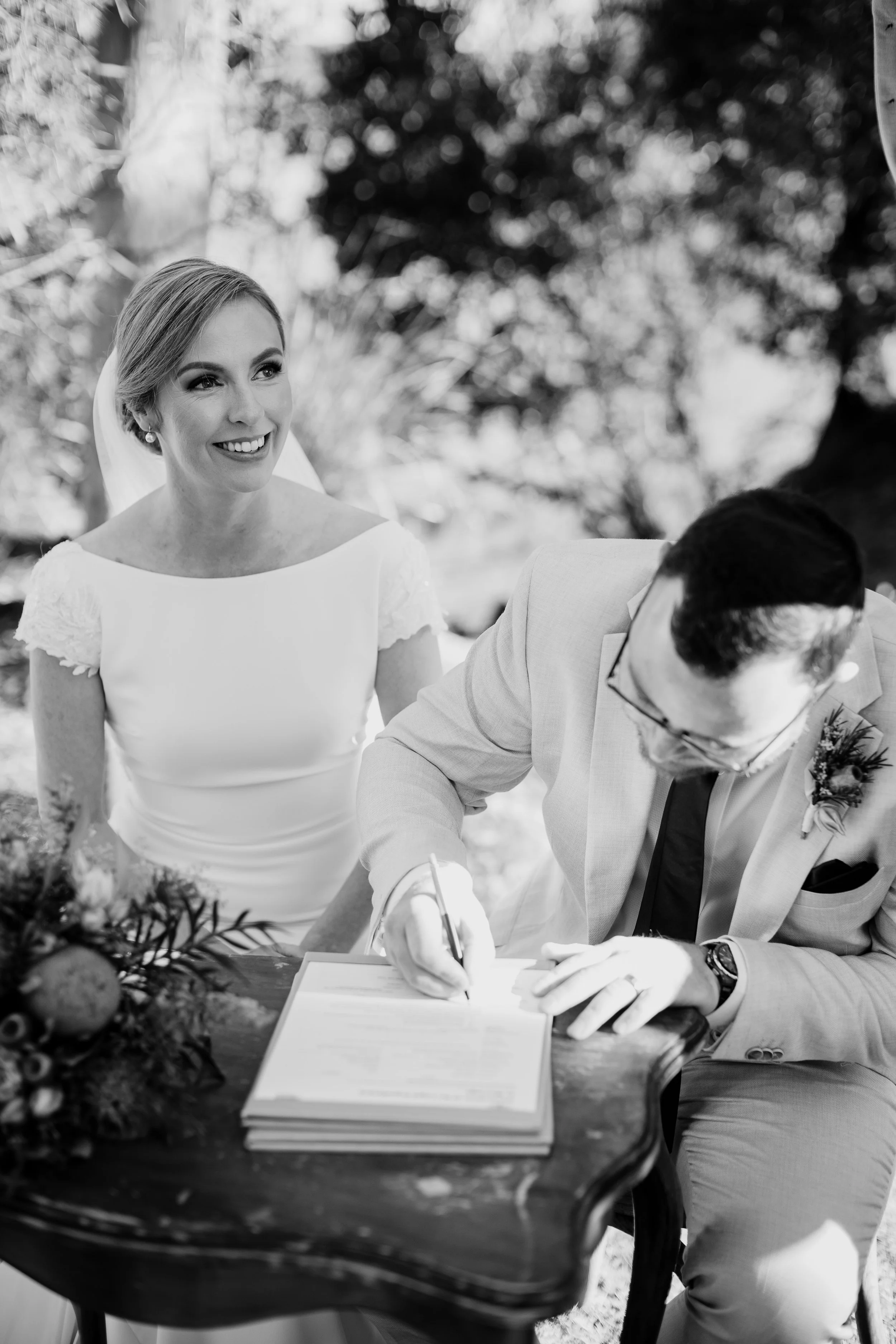 A bride smiling as a groom signs a marriage certificate during a wedding ceremony outdoors.