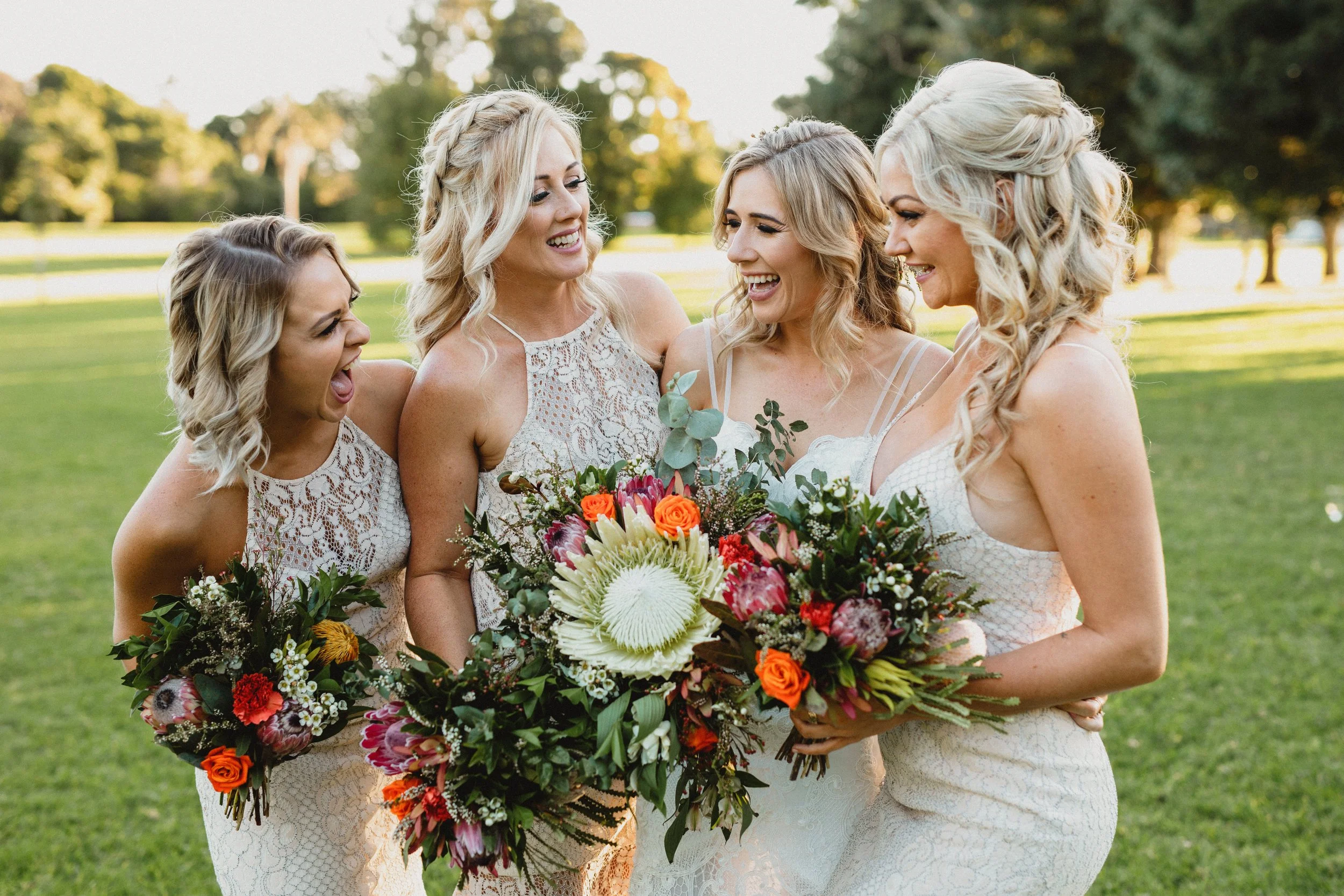 Five women in white dresses holding colorful bouquets, standing on a grassy field with trees in the background, laughing and celebrating.