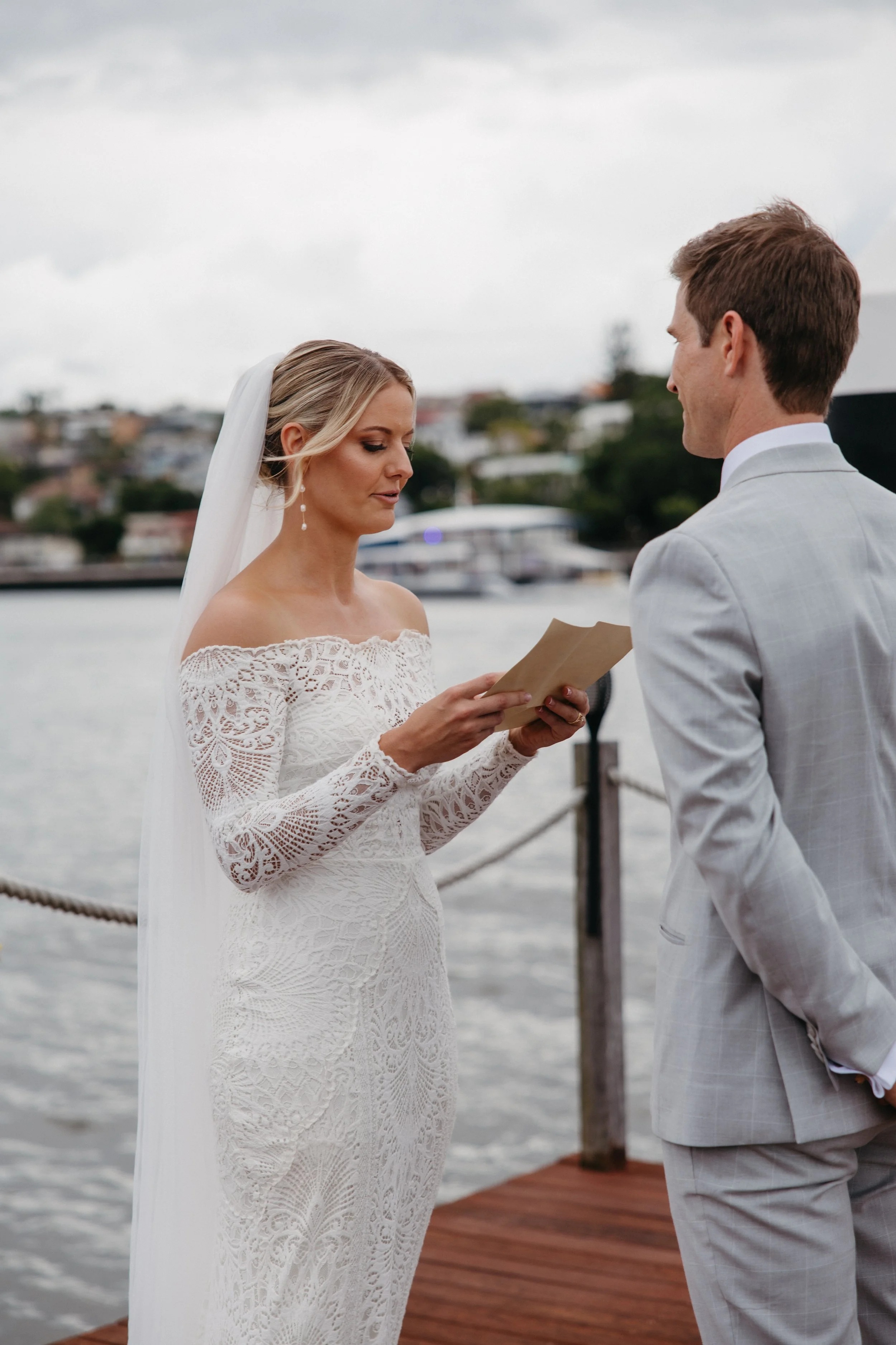 A bride in a white lace dress and veil reading vows to a groom in a light gray suit during a waterfront wedding ceremony.