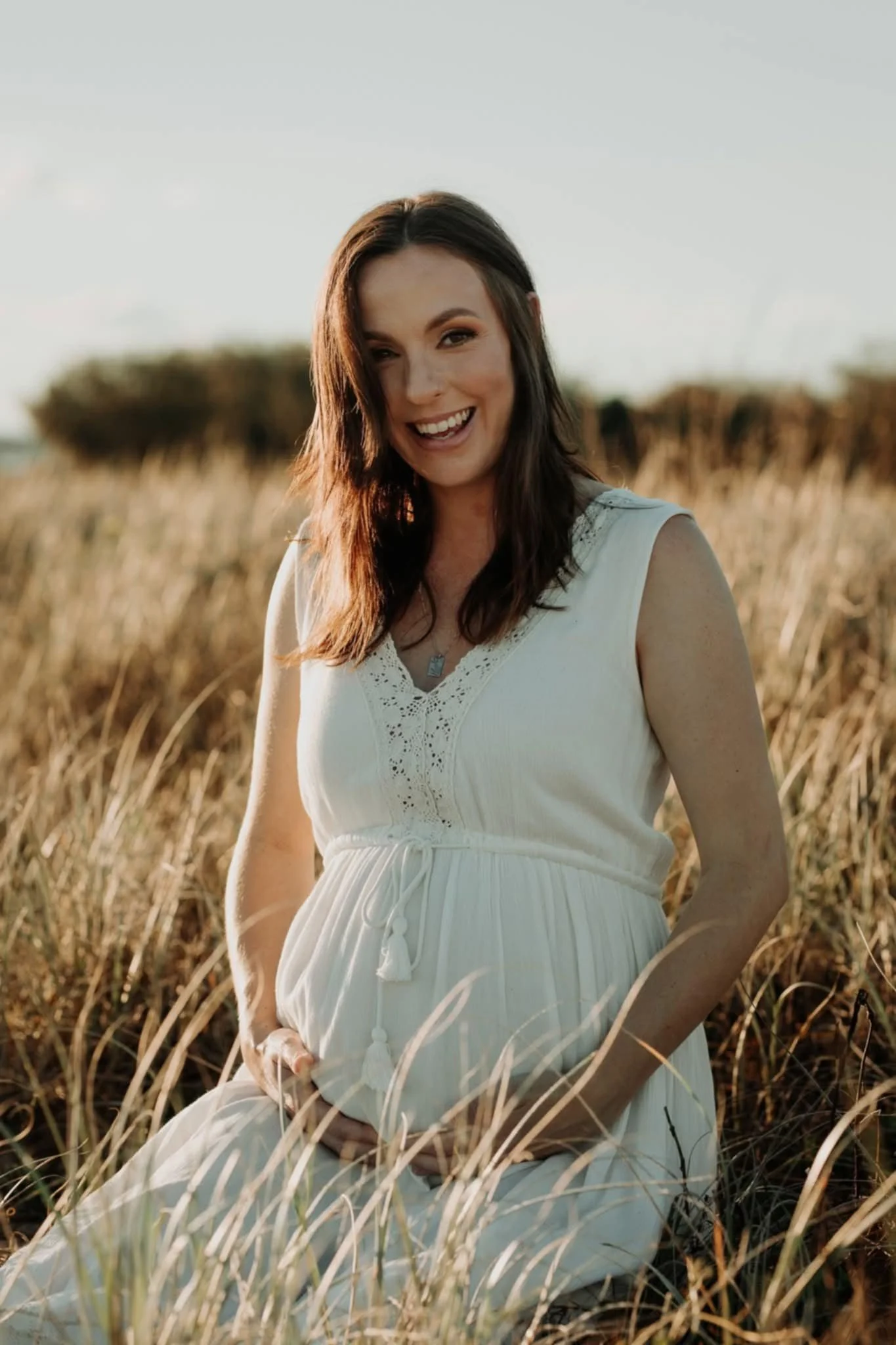 A smiling pregnant woman with long dark hair wearing a white sleeveless dress, sitting in a field of tall grass during golden hour sunlight.
