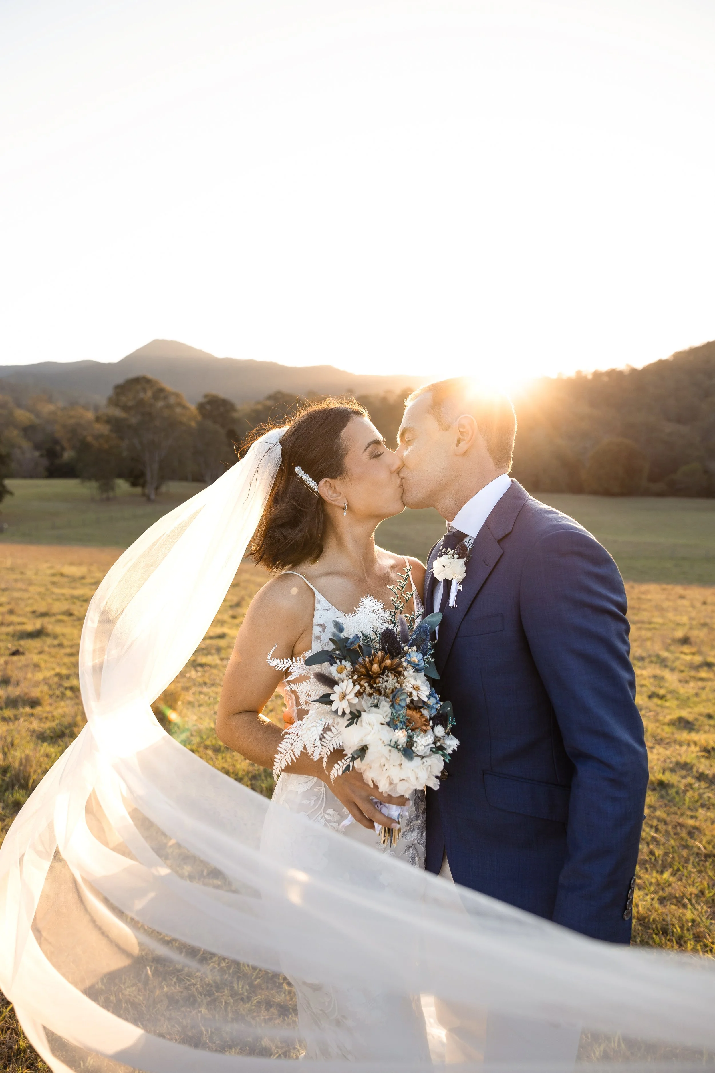 A bride and groom kissing outdoors at sunset, with a scenic landscape of mountains and trees in the background.
