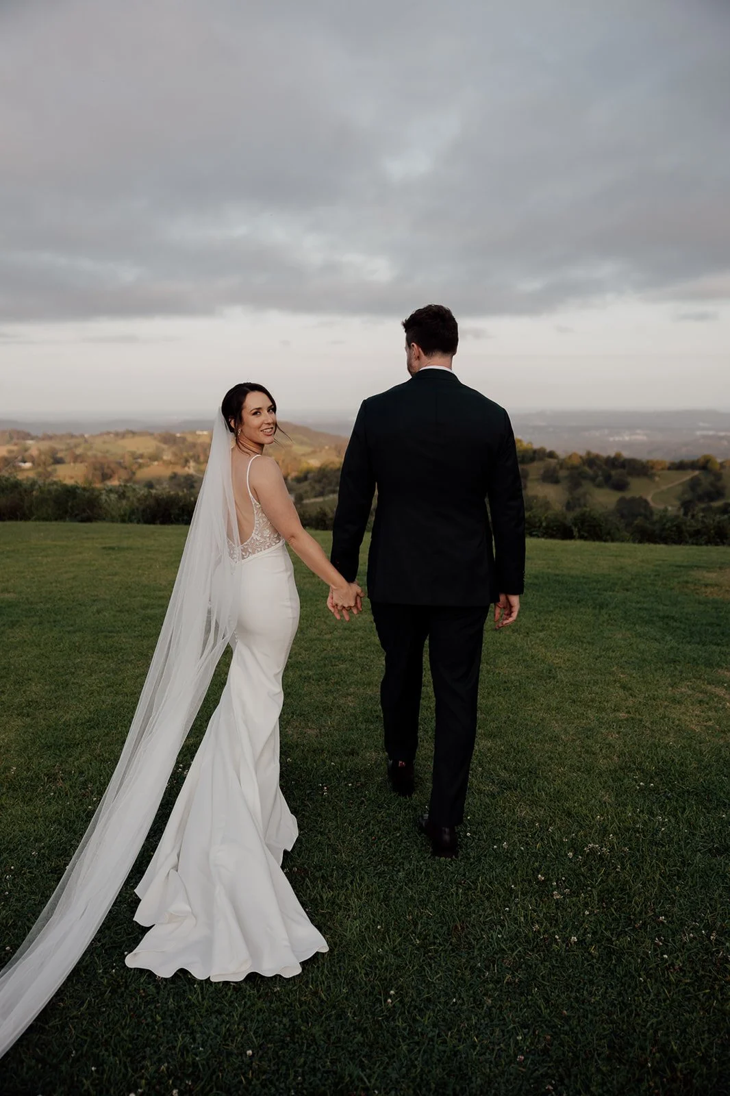 A bride and groom walking hand in hand across a grassy field with a scenic landscape in the background, under a cloudy sky.