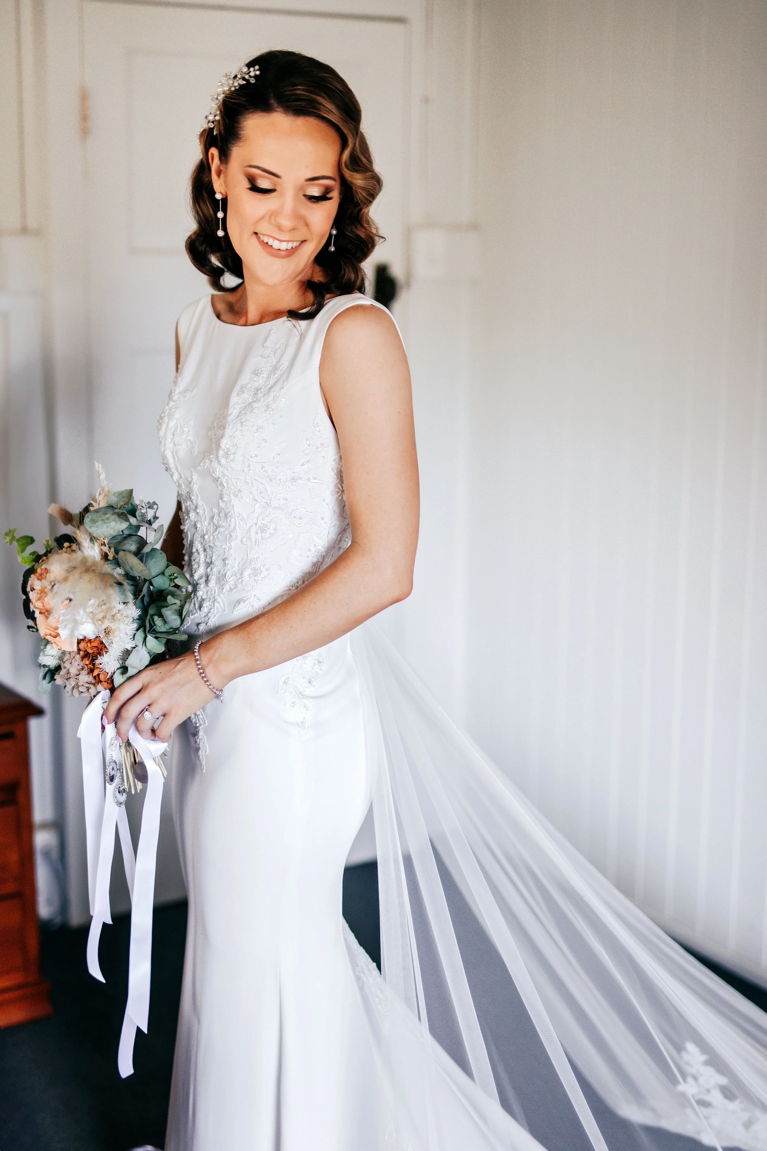 A bride in a white wedding dress holding a bouquet of flowers, smiling, with short dark hair styled in waves and wearing pearl earrings and a pearl bracelet, standing in a room with white walls.