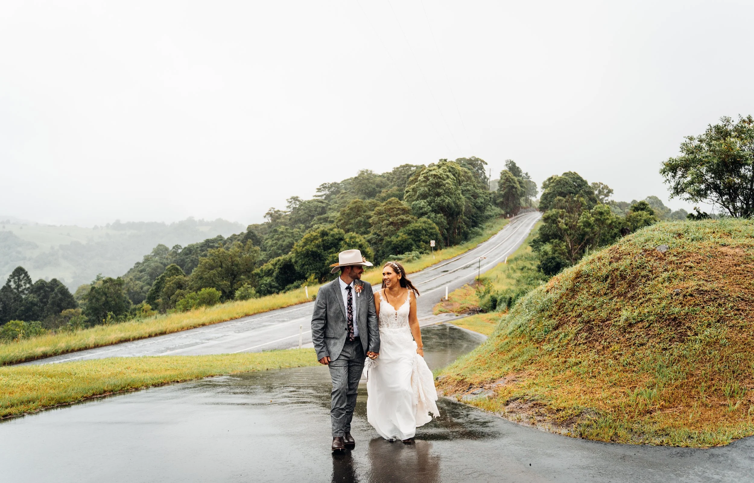 A couple in wedding attire walking on a wet road in a rainy, lush green countryside.