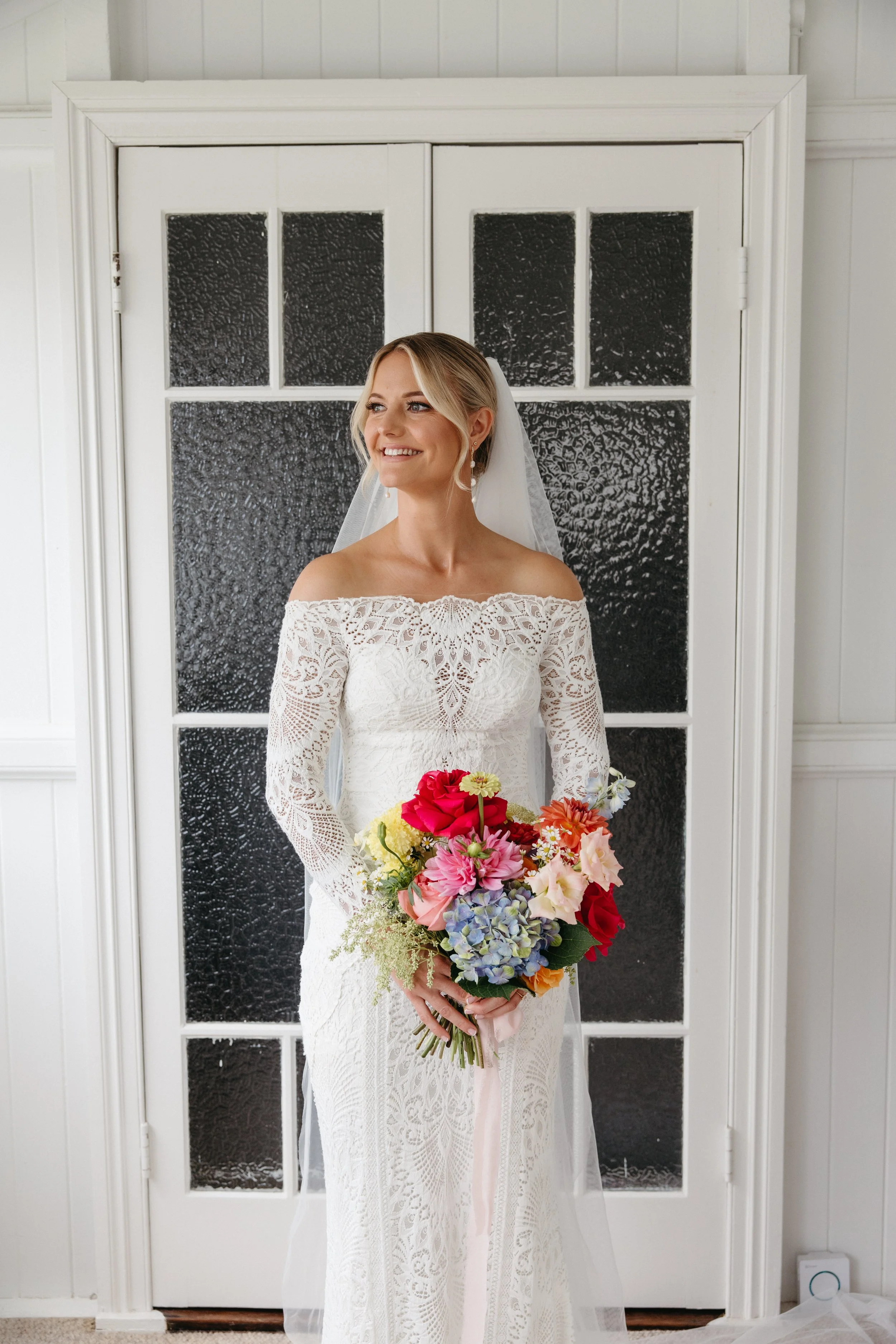 A bride in a white lace off-the-shoulder wedding dress holding a colorful bouquet of flowers, standing in front of a white door with textured glass panels, smiling.
