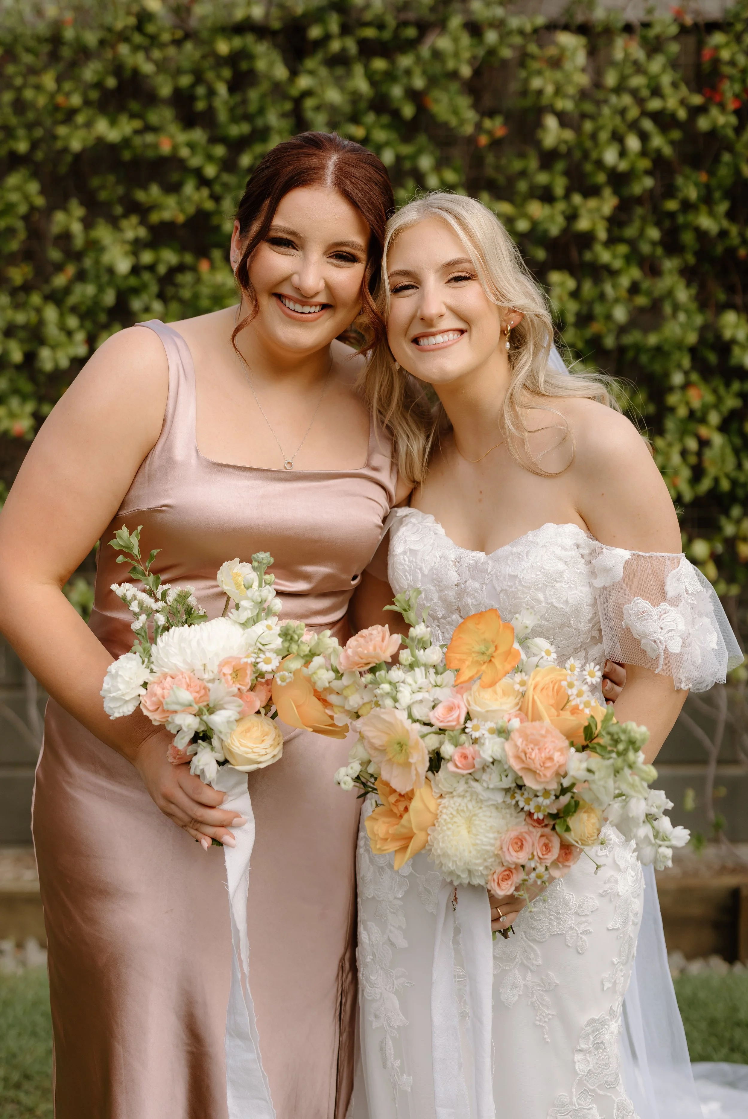 Two women at a wedding, one woman in a white lace wedding dress holding a bouquet and the other in a bedazzled soft pink dress holding a bouquet, standing outside against a background of greenery with small pink and white flowers, smiling at the came