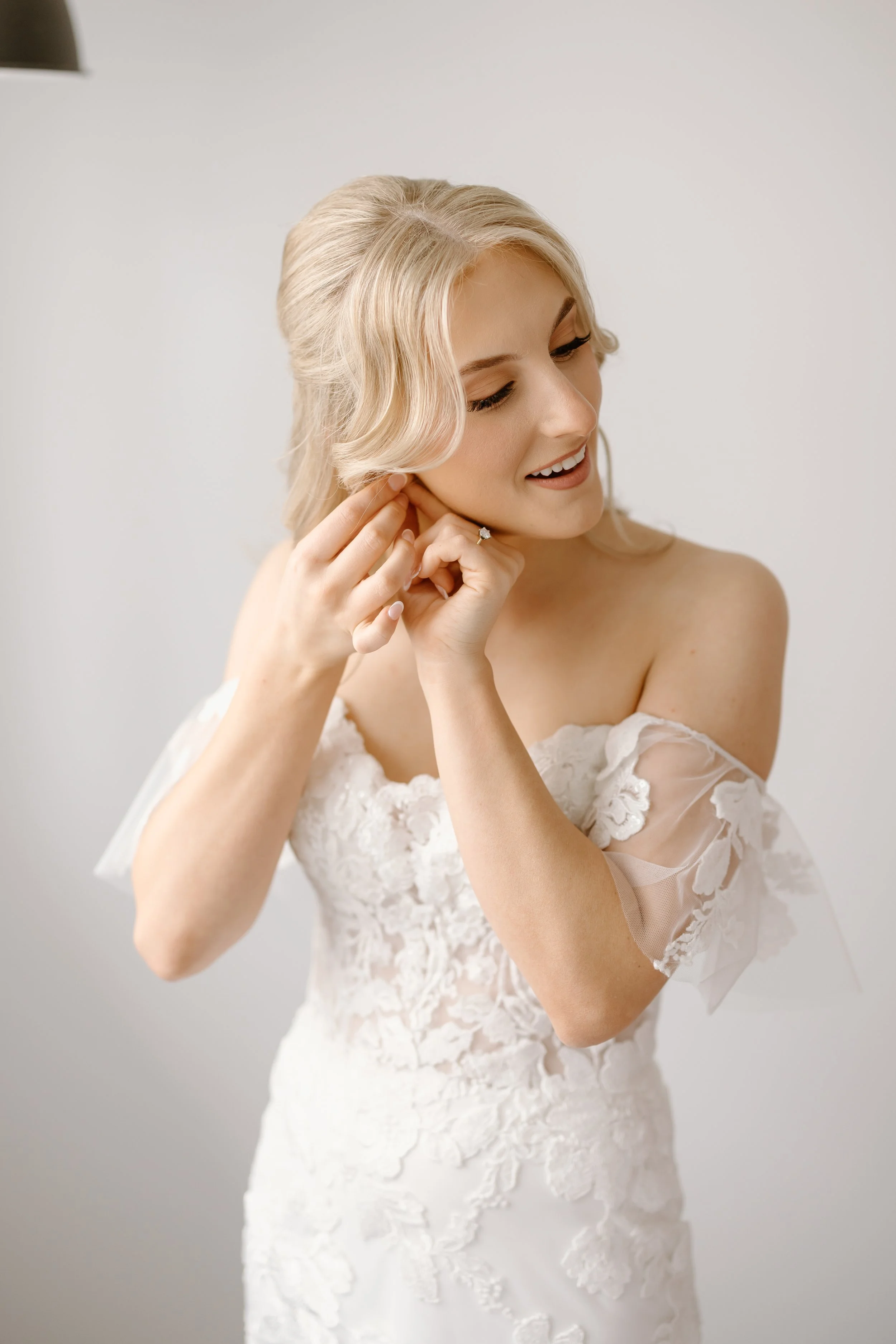 A bride with blonde hair in an updo, wearing a white lace wedding dress, adjusts her earring while smiling gently.