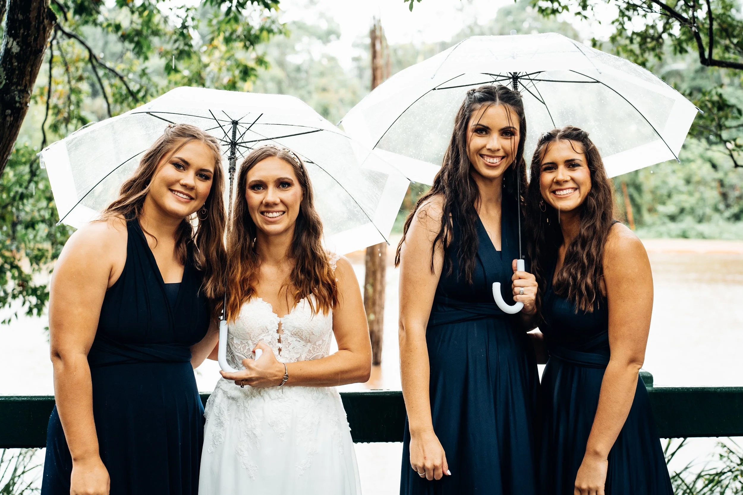 Four women standing outdoors in the rain, holding umbrellas, smiling for a photo. One woman in white appears to be a bride, and the others are bridesmaids dressed in dark dresses.