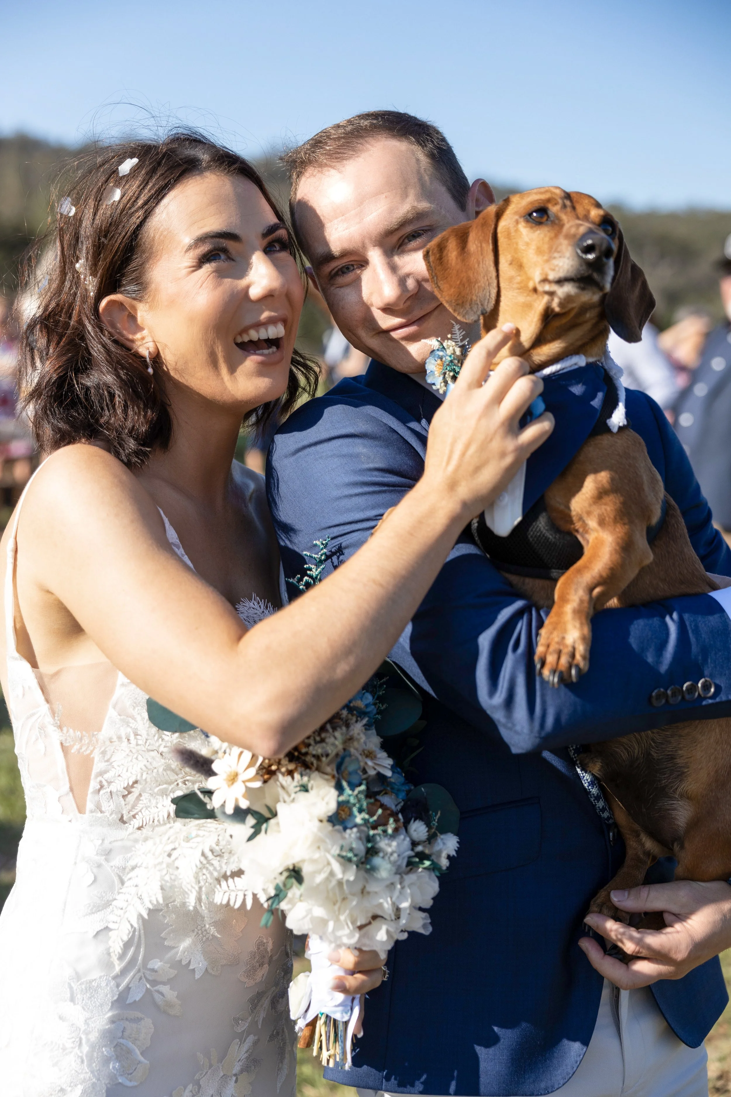 A bride and groom holding a dachshund dog at their wedding celebration outdoors.