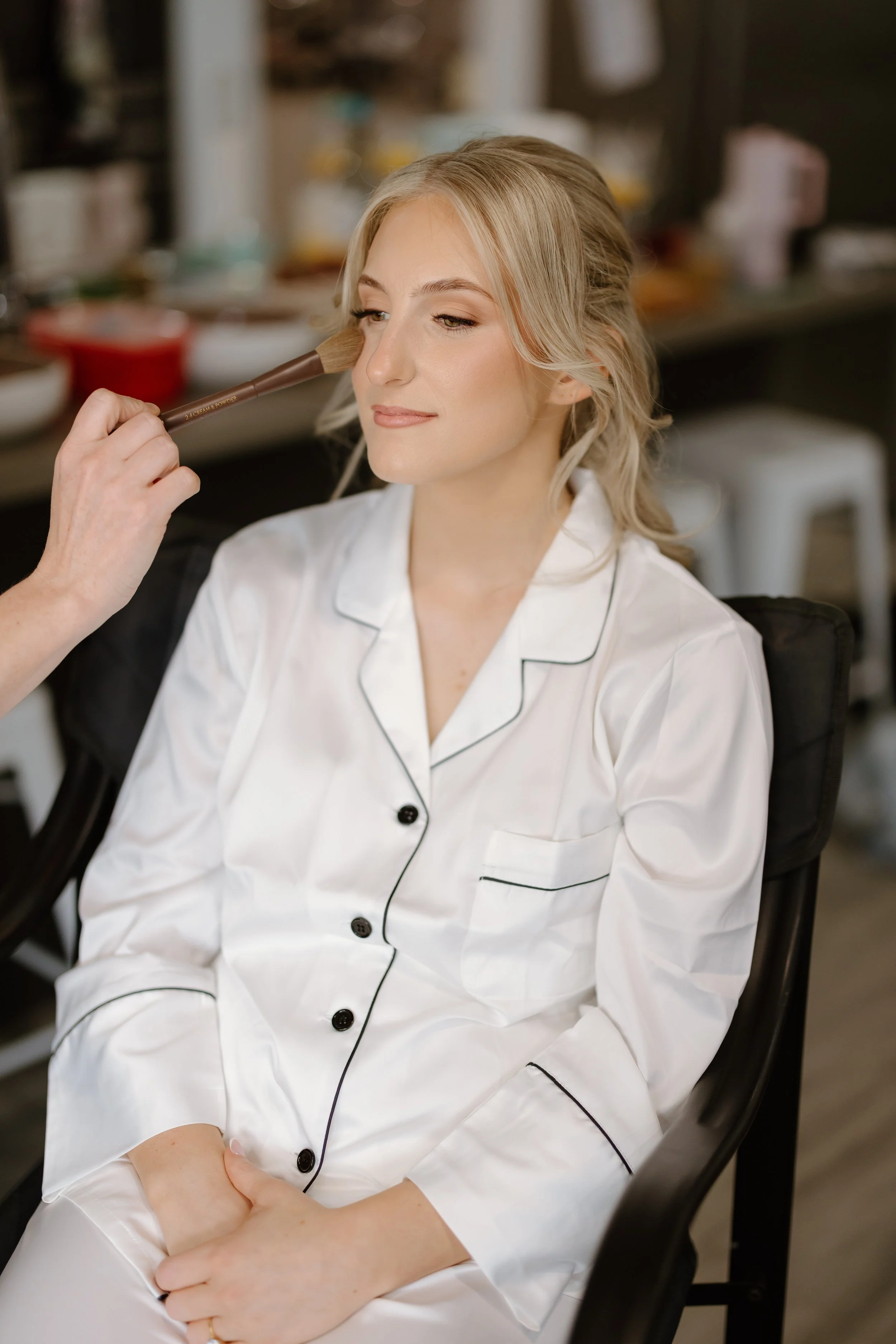 A woman in white pajamas sitting in a chair with her eyes closed as makeup artist applies blush with a brush on her cheek in a well-lit room.