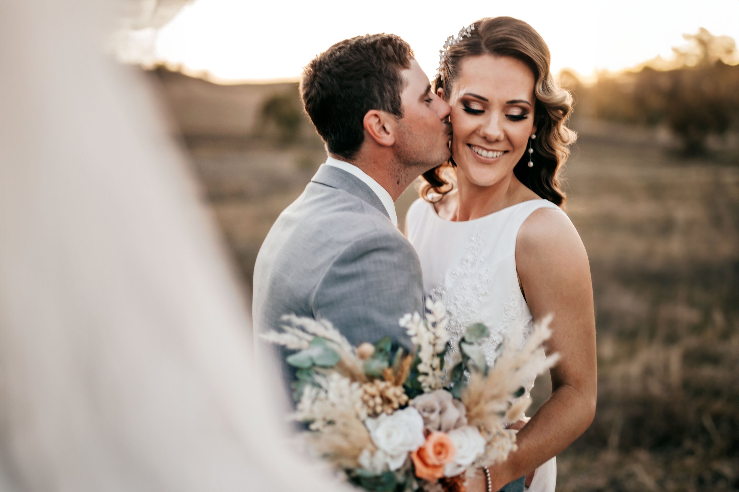 A newlywed couple in wedding attire, with the groom kissing the bride's cheek as she smiles, holding a bouquet with white and peach flowers, outdoors during sunset.