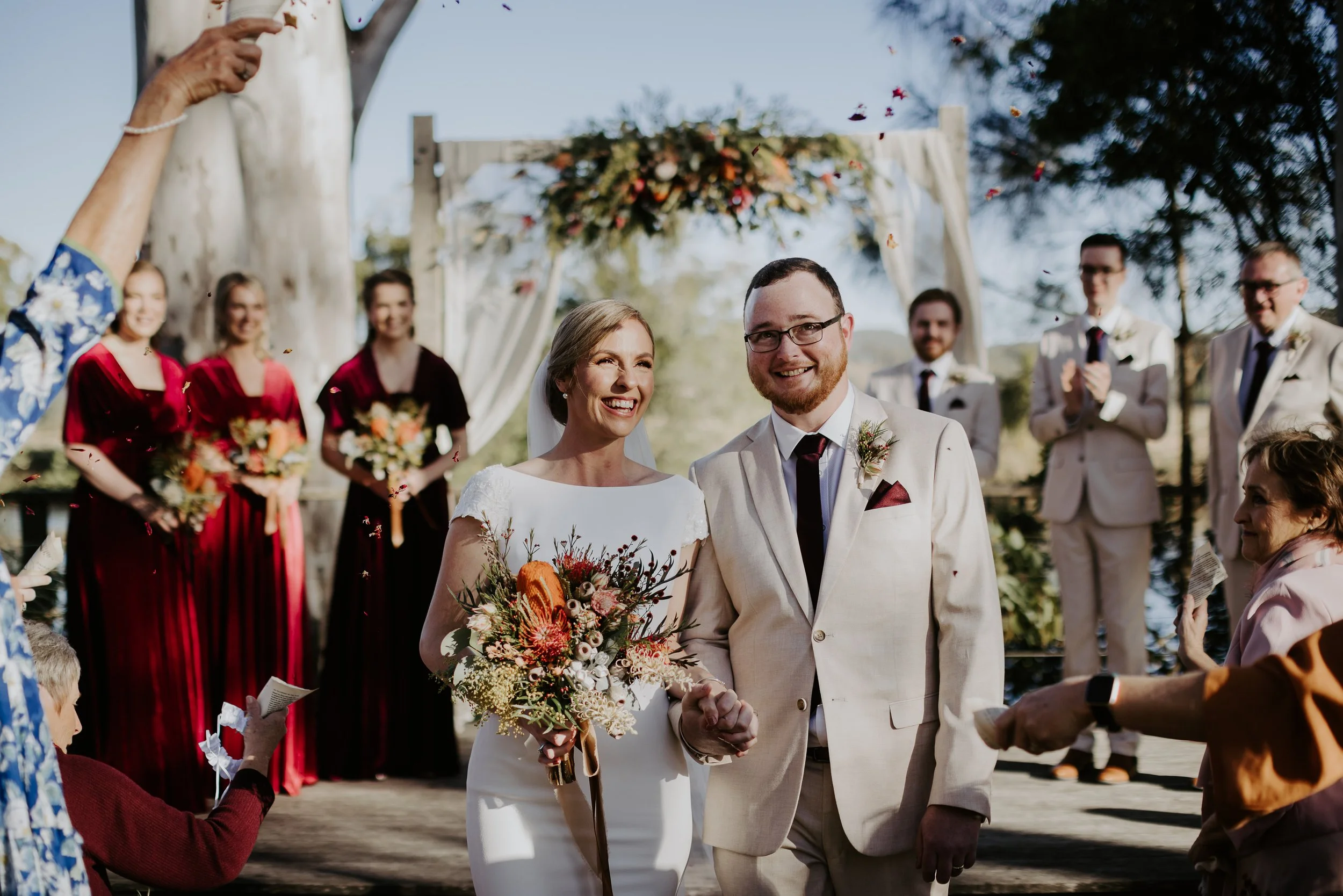 Bride and groom smiling and holding hands during outdoor wedding ceremony, surrounded by bridal party and guests, with confetti in the air, trees, and sky in the background.