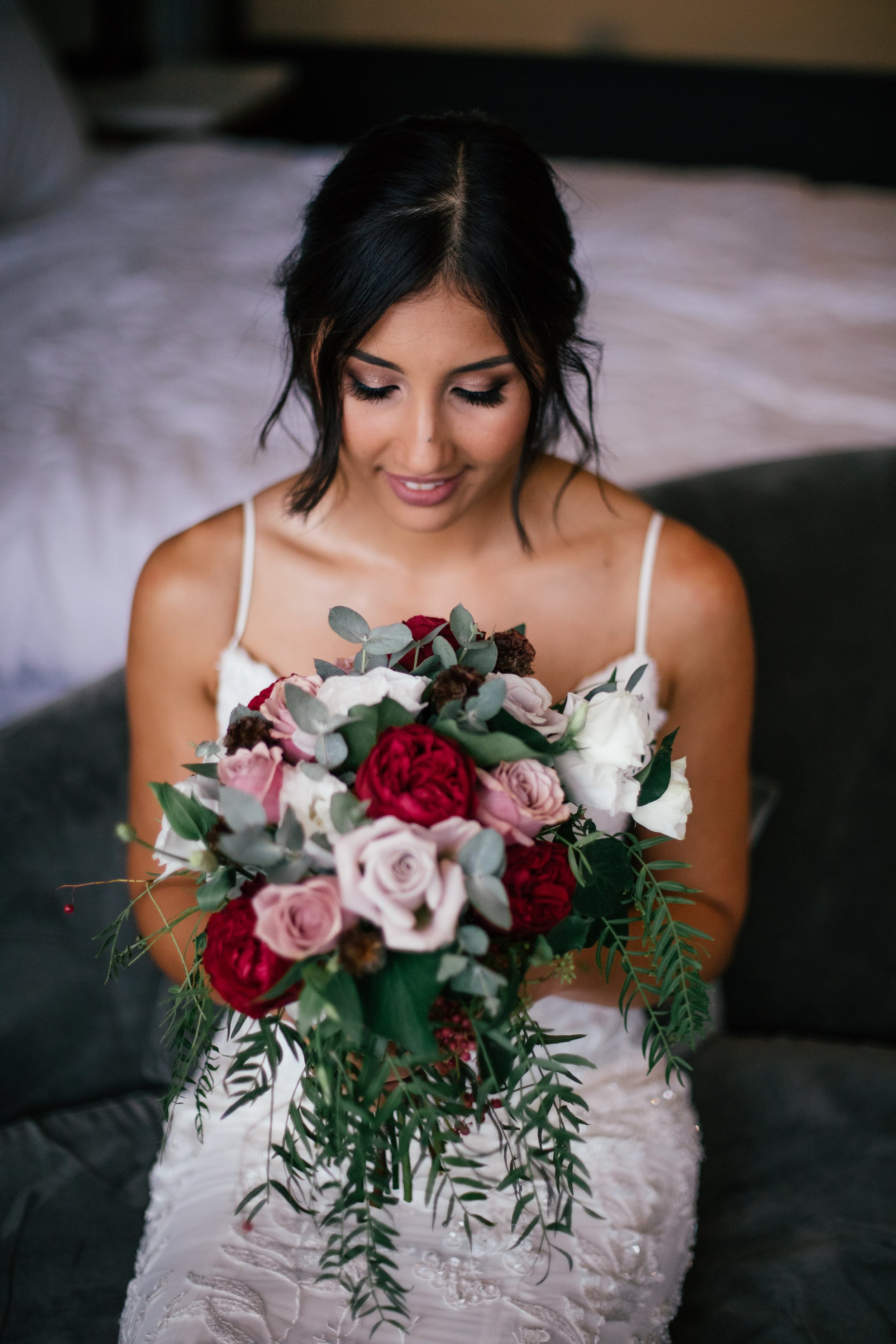 A woman in a wedding dress holding a bouquet of red, pink, and white flowers, sitting on a dark sofa.
