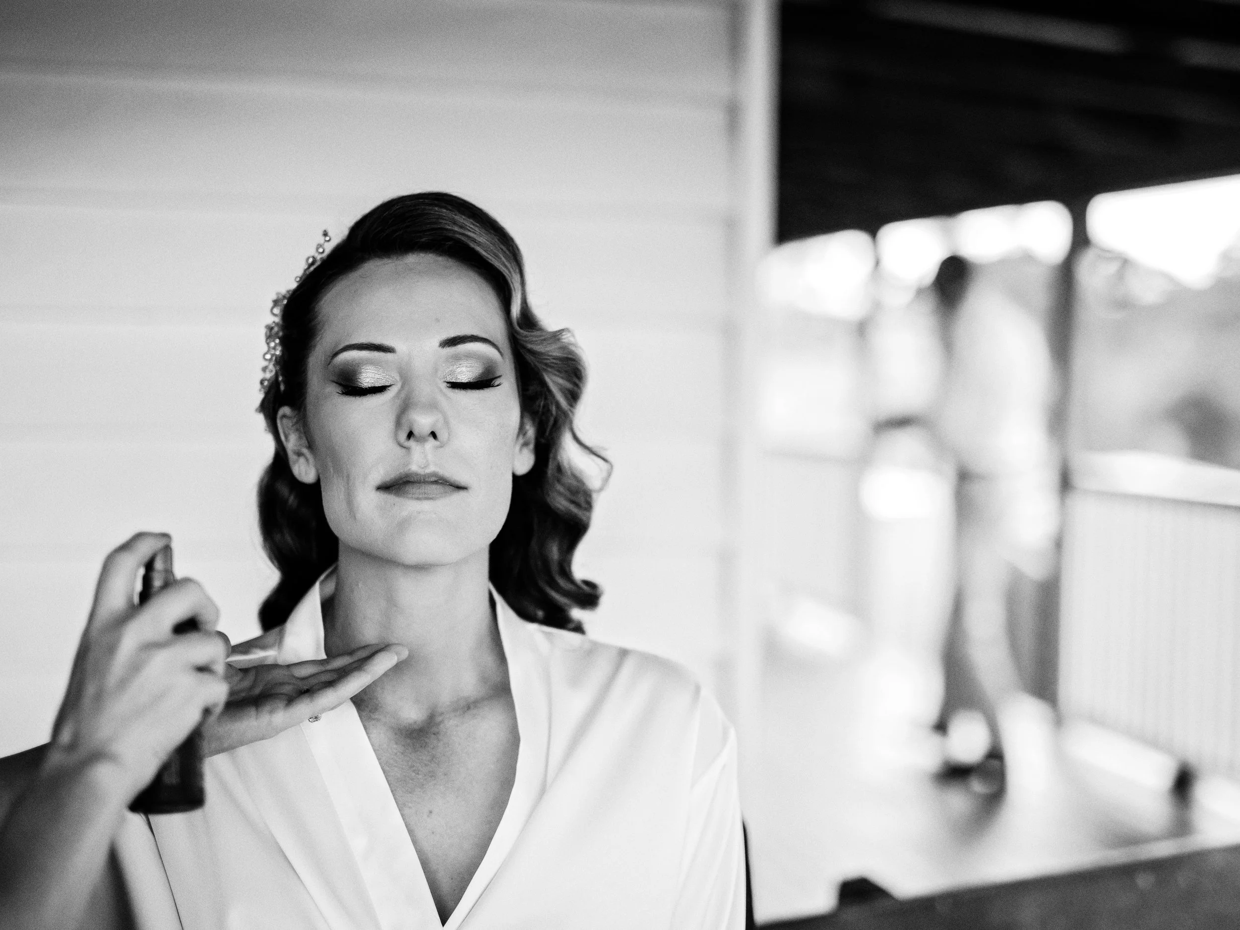 A woman with styled hair and makeup has her eyes closed as a makeup artist sprays her face with a spray bottle. She is wearing a white robe, and the background shows a porch or balcony with blurry figures walking in the distance.