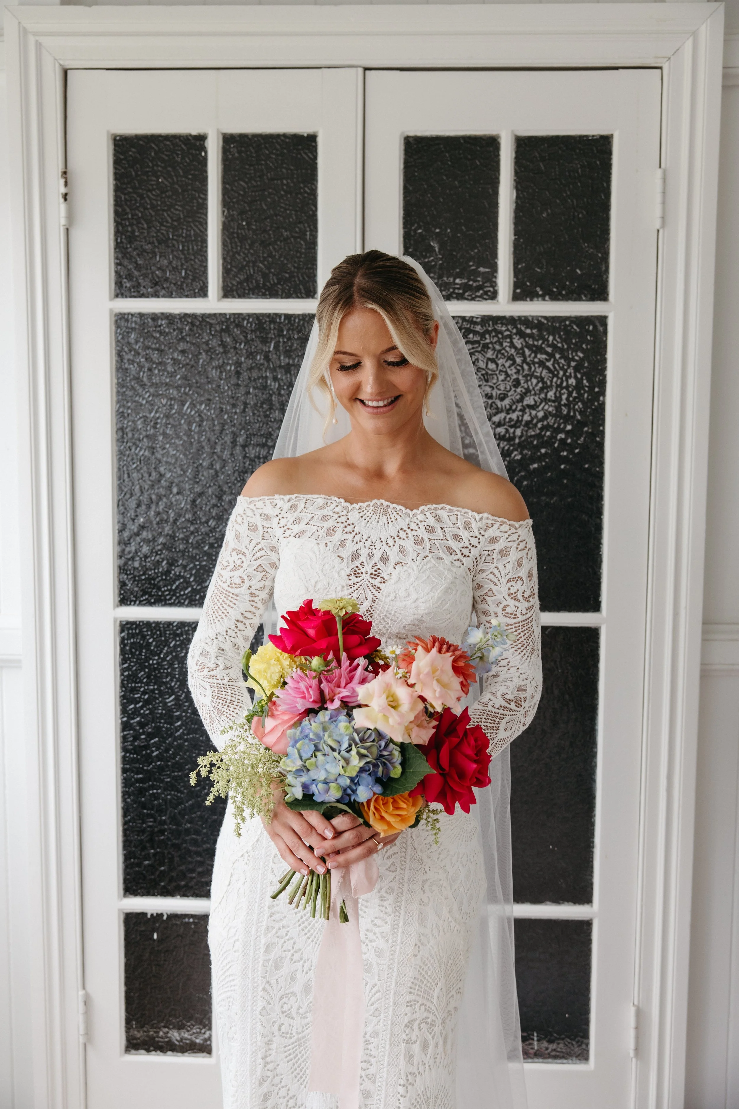 A bride in a white lace off-shoulder wedding dress holding a colorful bouquet of flowers standing in front of a window with textured glass panes.