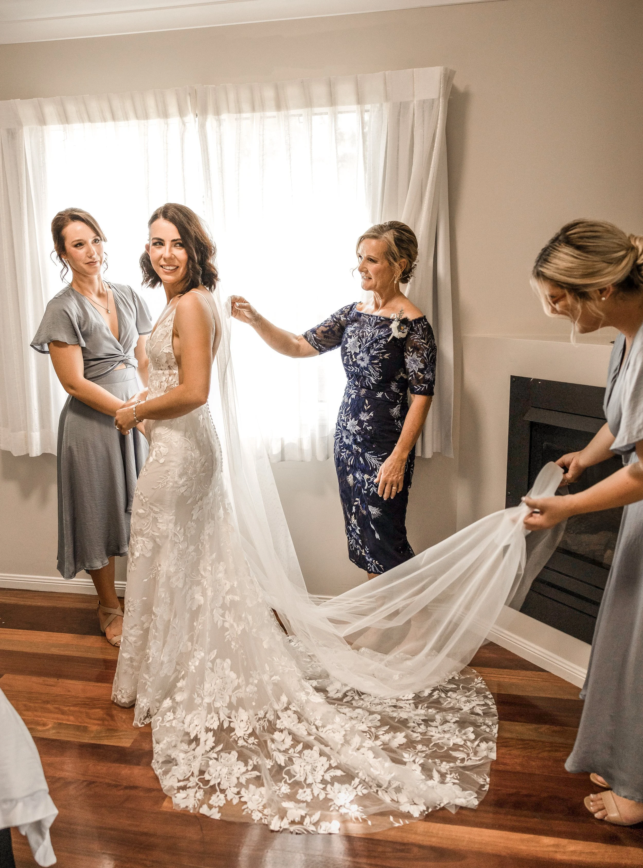 A bride in a lace wedding dress standing with three women, while two women assist with her long train and the bride's mother looks on, in a room with a window and white curtains.