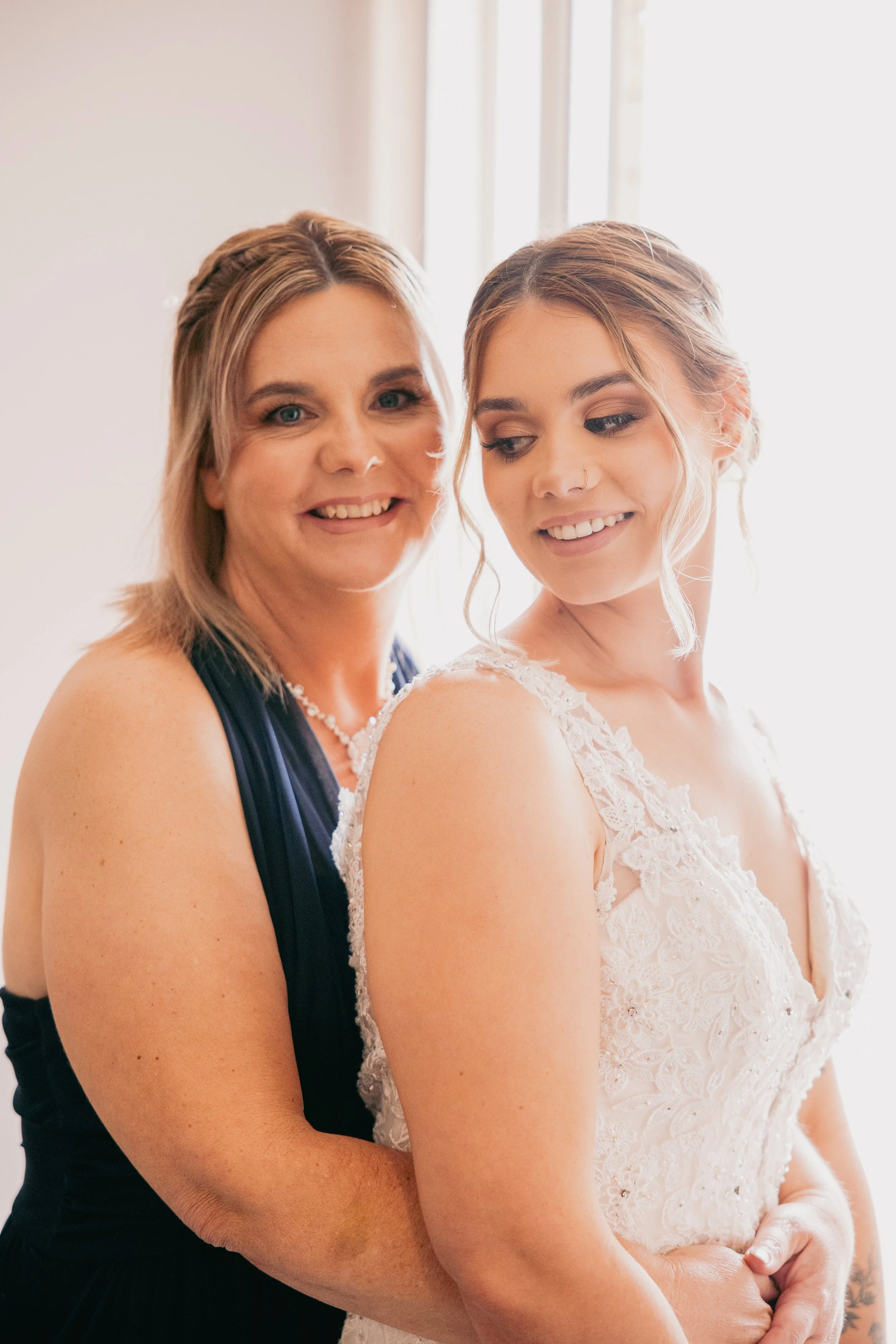 A woman in a navy dress with pearls is hugging a bride in a white lace wedding dress, both smiling softly, standing by a window with natural light in the background.