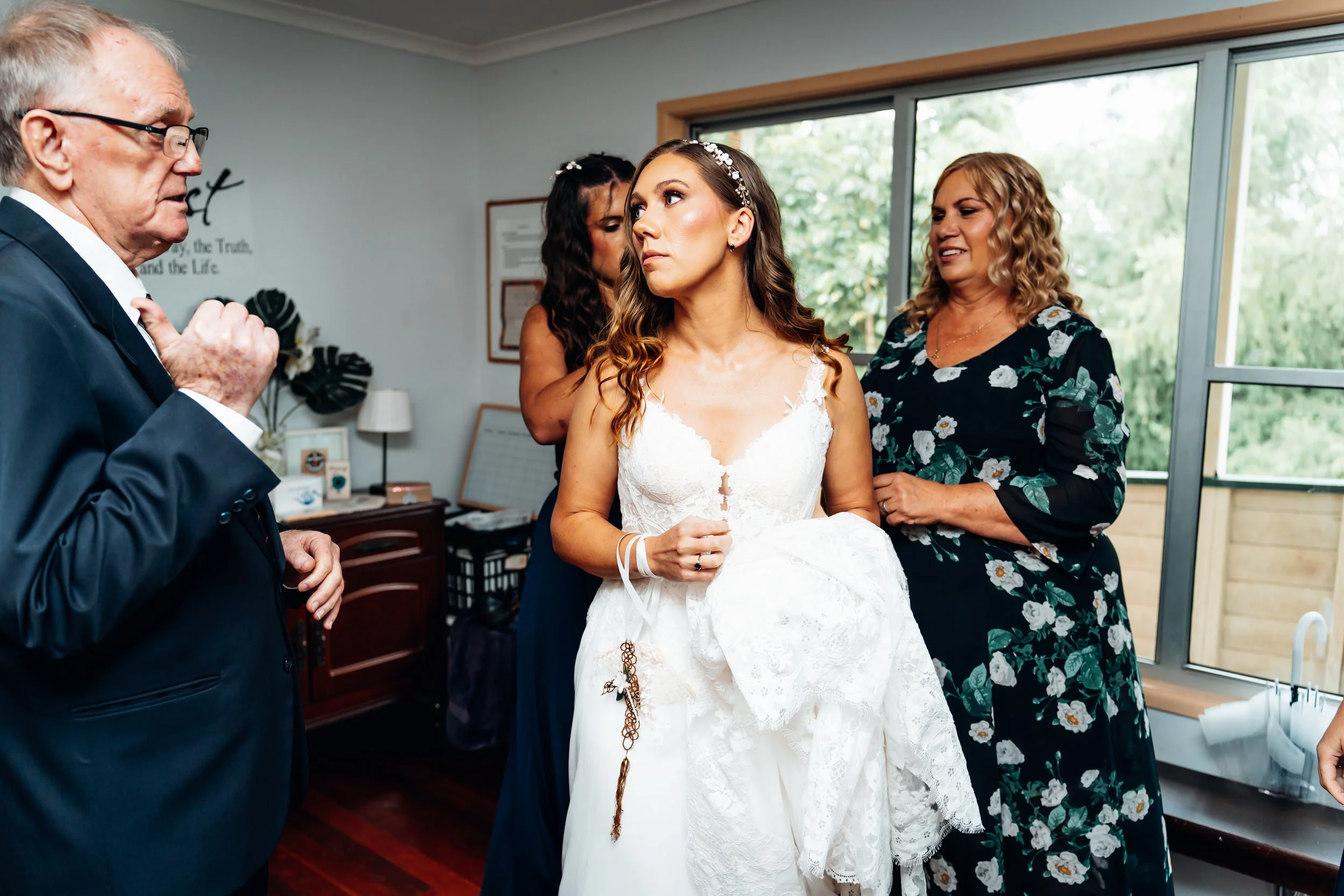 A bride in a white wedding dress holding her shoes while her family helps her prepare for her wedding.