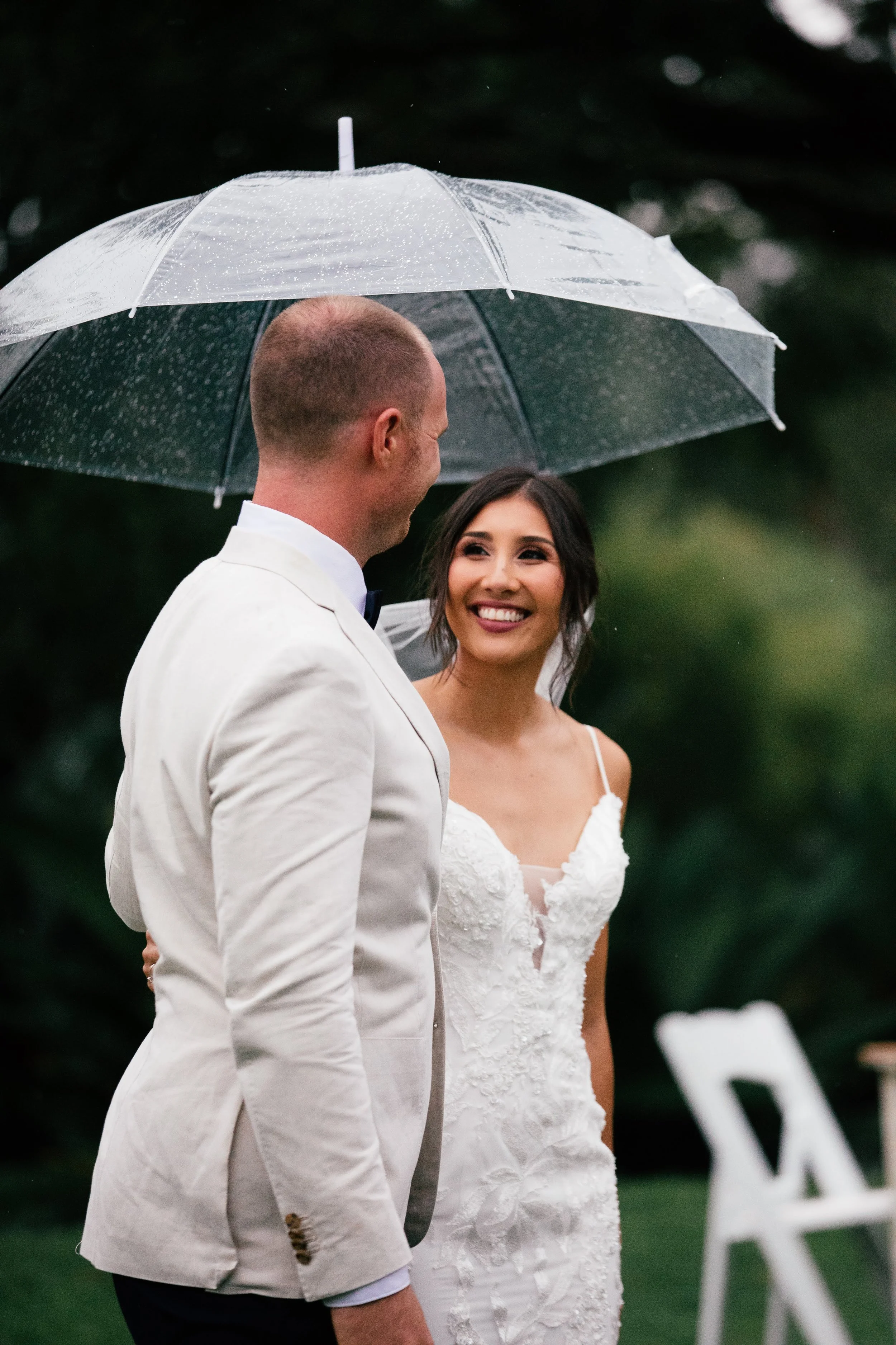 A bride and groom sharing a moment under a clear umbrella during their wedding outdoors in the rain, with the bride smiling warmly at the groom.