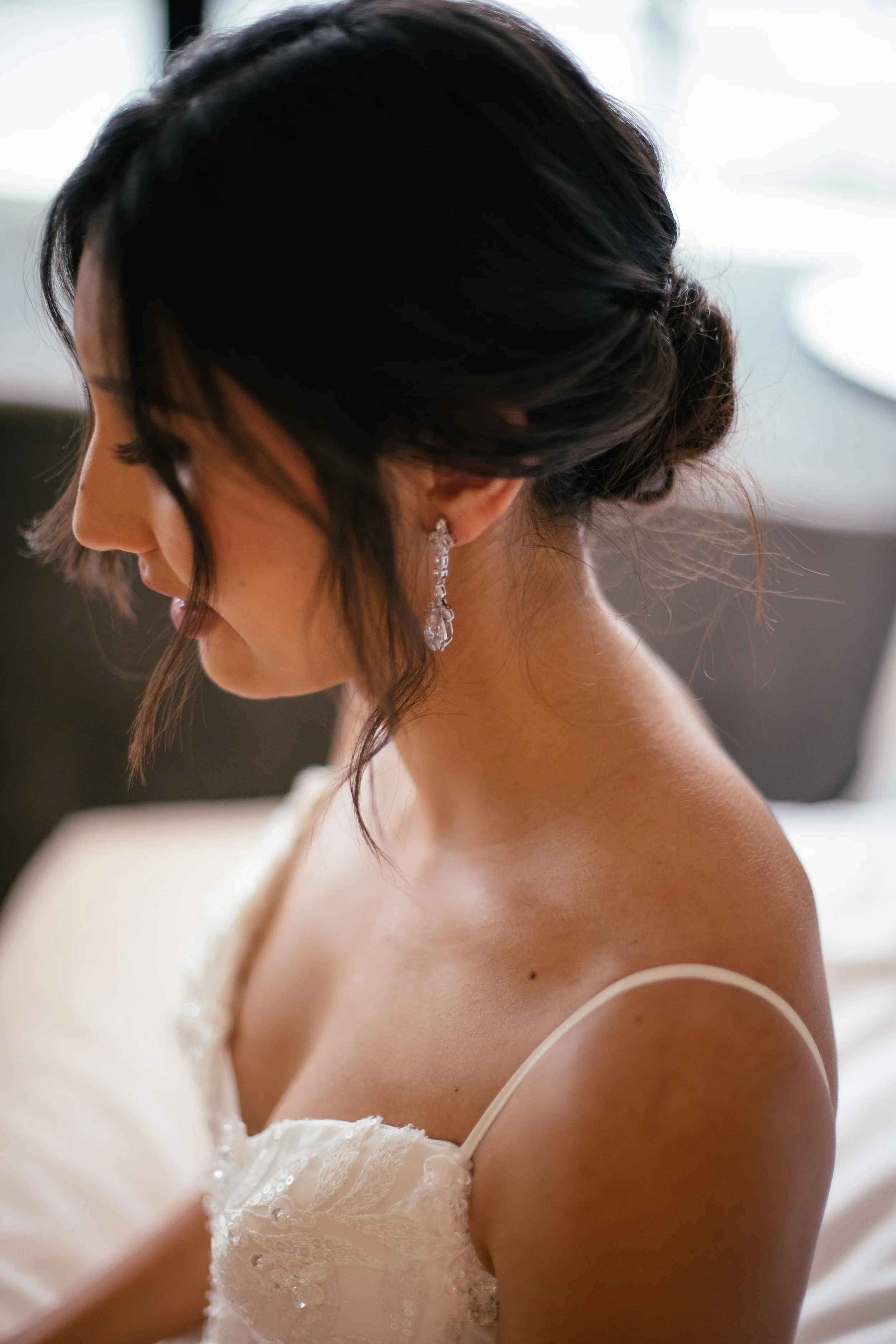 Close-up of a bride with dark hair styled in an elegant updo, wearing diamond earrings and a off-shoulder lace wedding dress, looking down thoughtfully.