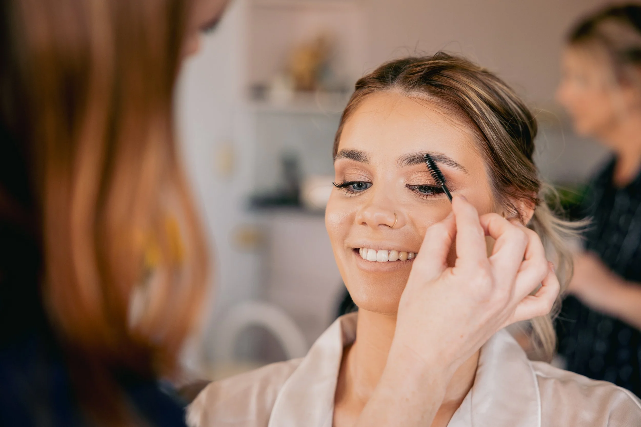 A makeup artist applying eyebrow makeup to a smiling woman while another person is blurred in the background.