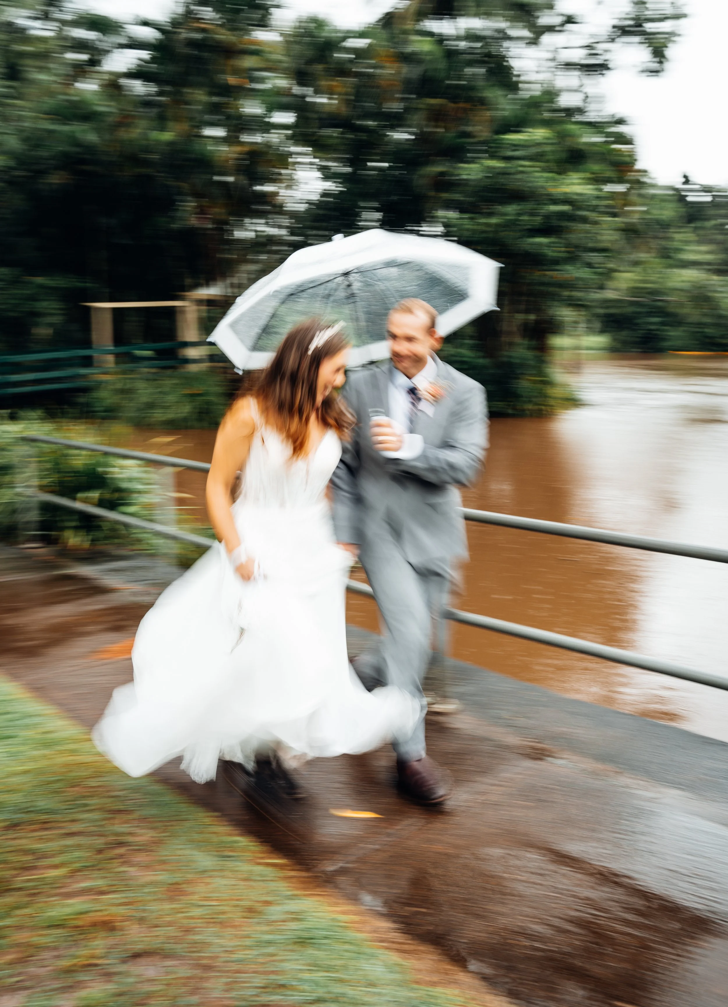 A bride and groom walking briskly in the rain under an umbrella near a river, with wedding attire and wet surroundings.