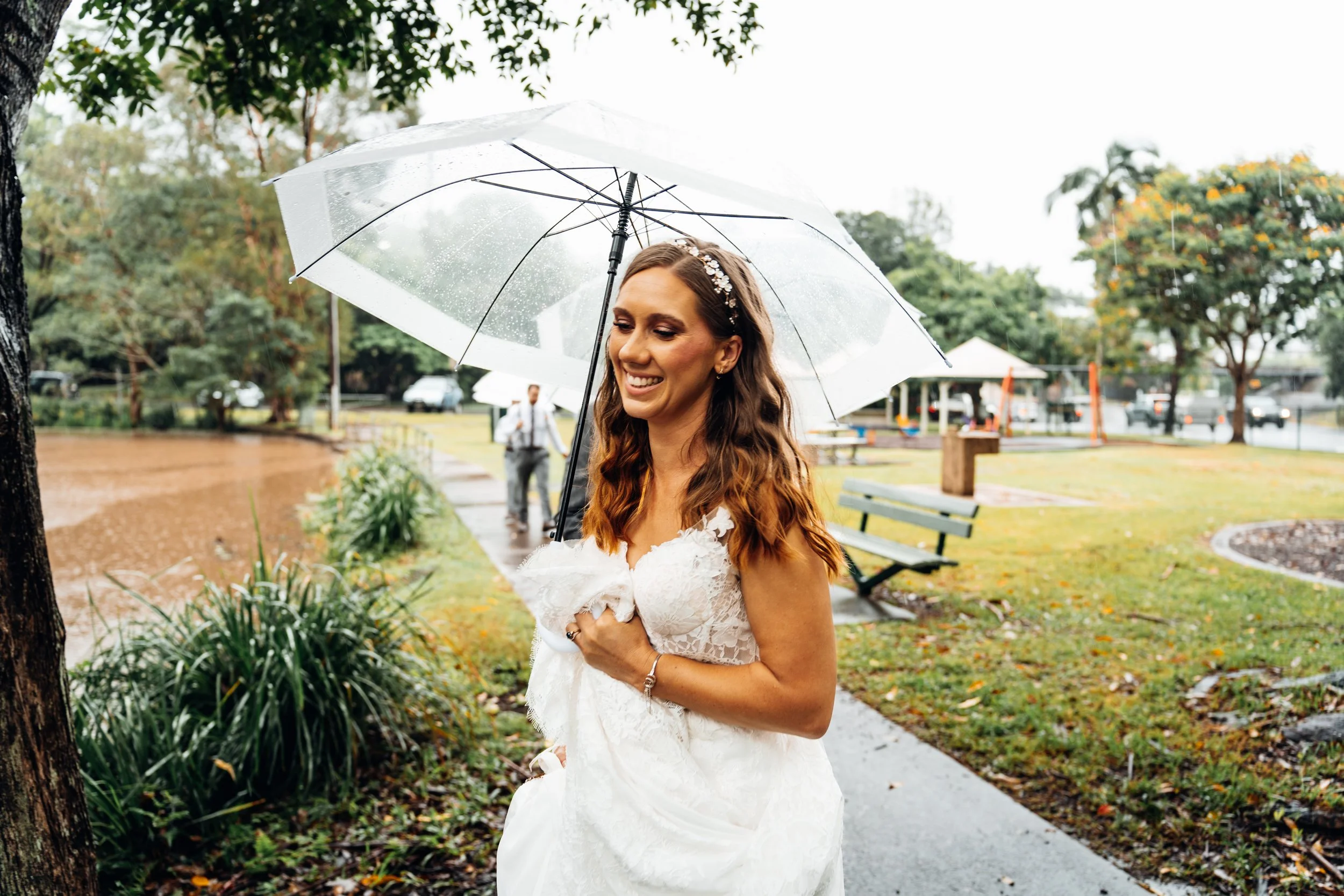 A bride in a white wedding dress holding an umbrella on a rainy day in a park.