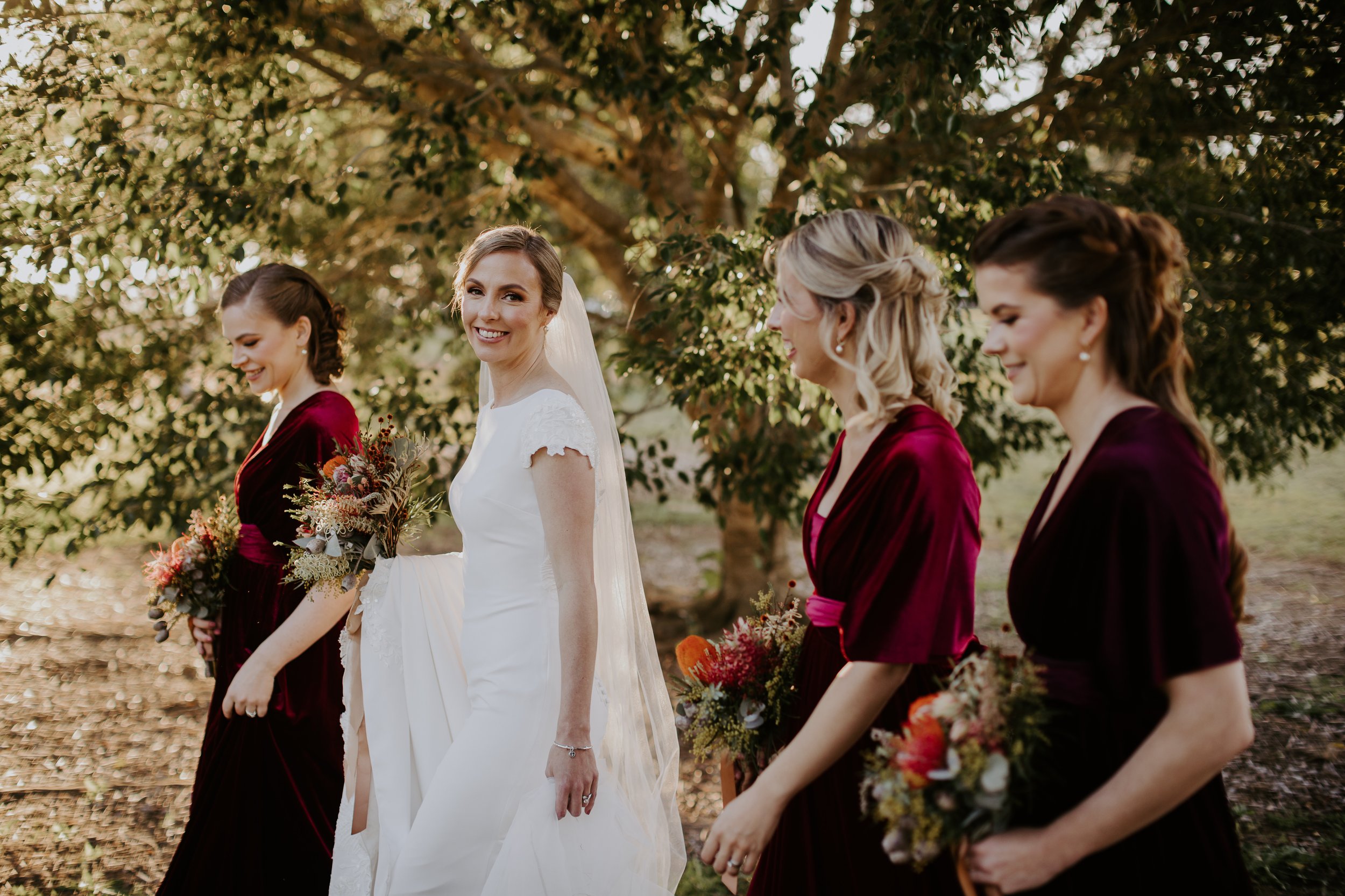 A bride in a white wedding dress and four bridesmaids in maroon dresses holding bouquets, walking outdoors near a large tree.