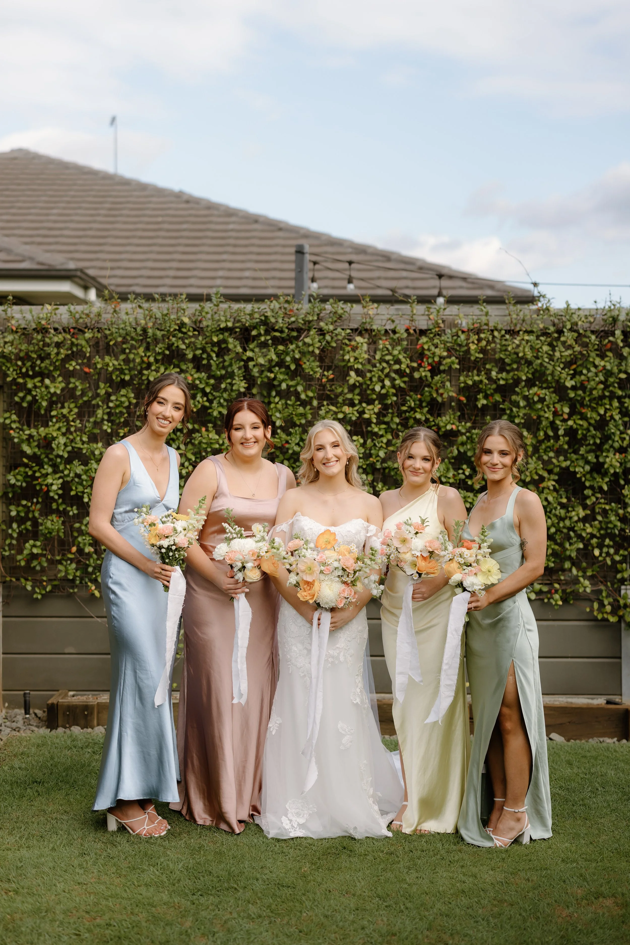 Five women, including a bride in a white wedding dress, standing outdoors on grass with a green leafy hedge background, holding bouquets of flowers.