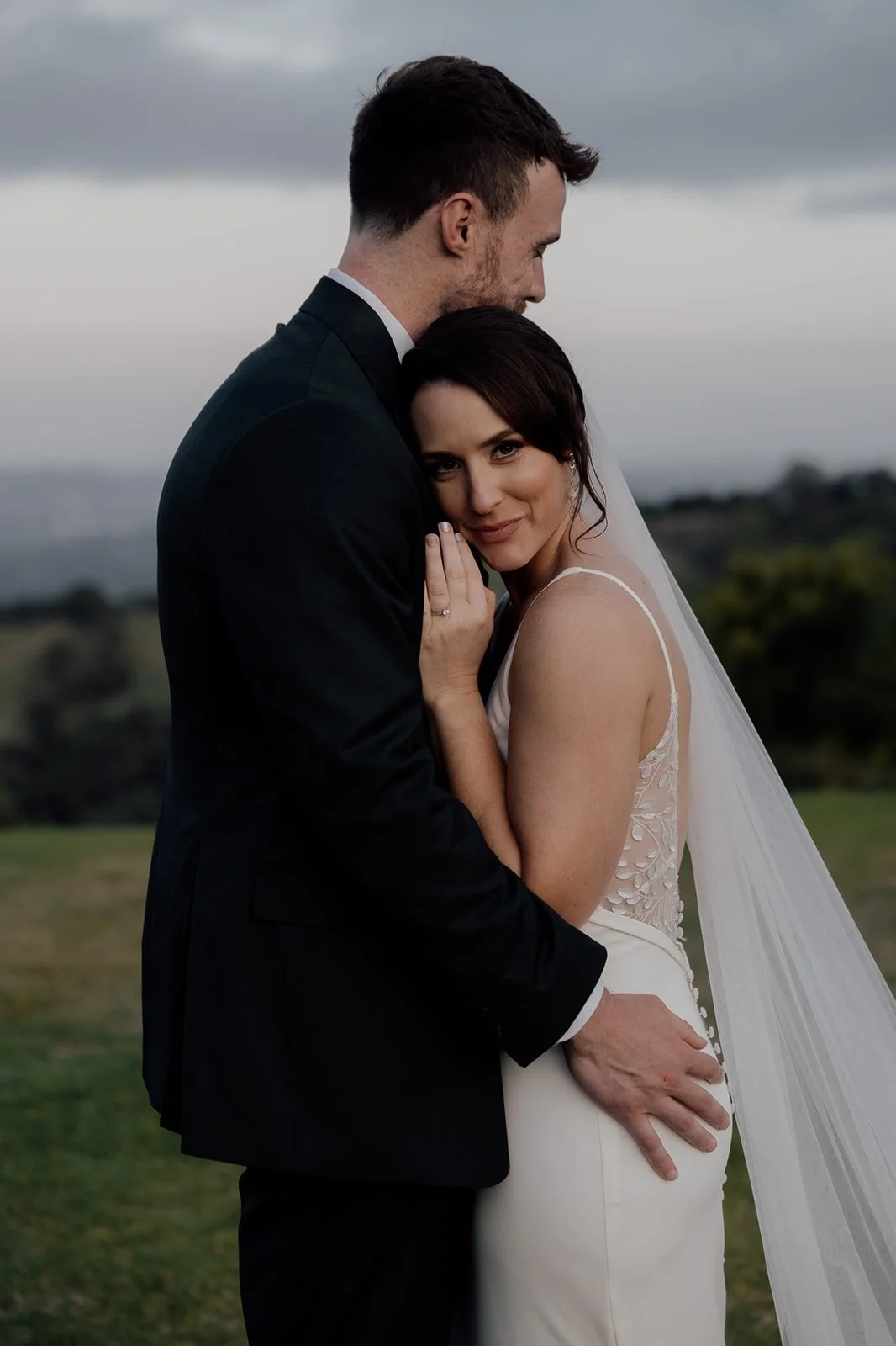 A bride and groom sharing an intimate moment outdoors, with the bride smiling and leaning into the groom, who is gently holding her waist. The woman is wearing a white wedding dress with a veil, and the man is in a black tuxedo.
