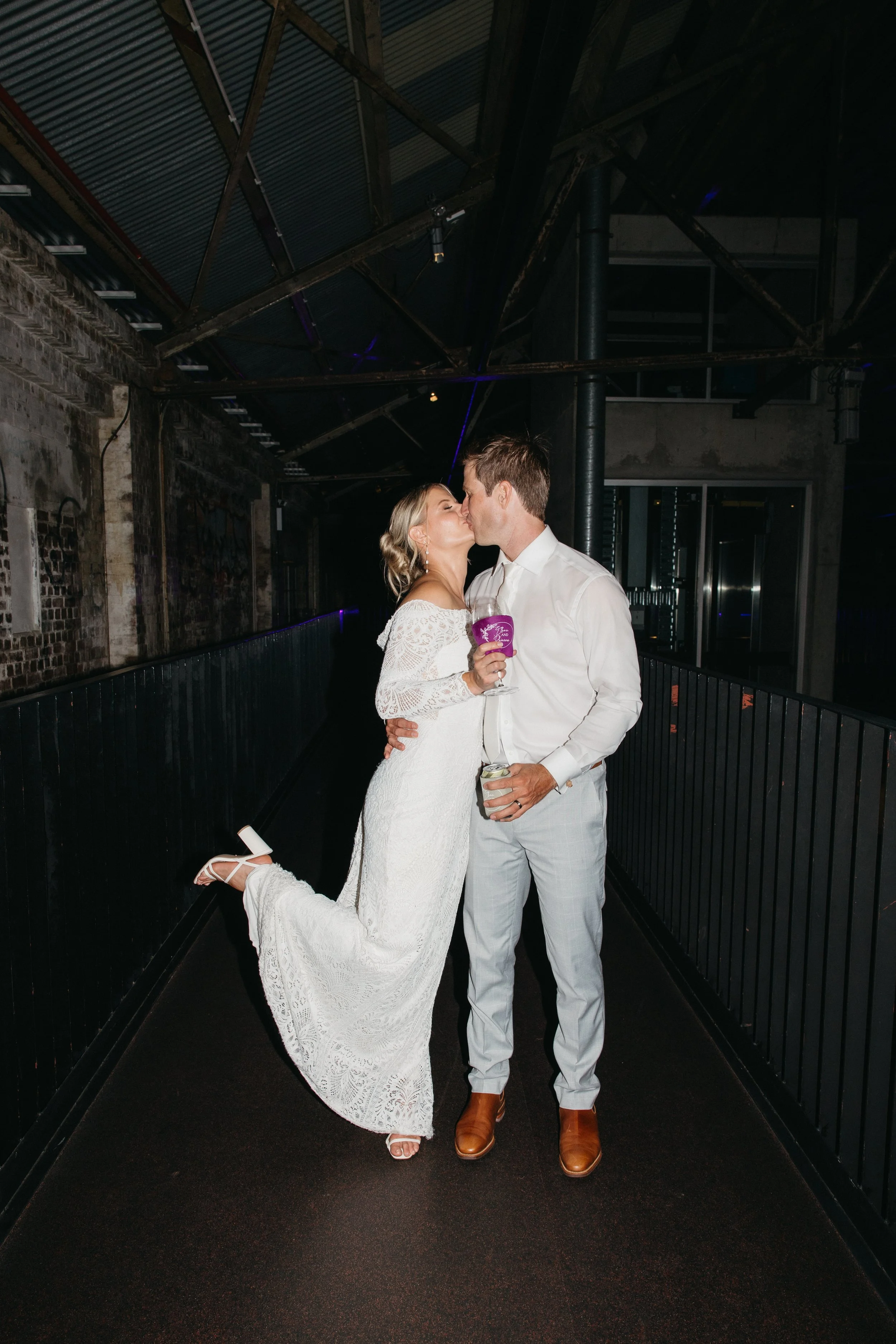 A bride and groom sharing a kiss at their wedding reception. The bride is wearing a white lace wedding dress and the groom is dressed in light gray pants and a white shirt. They are standing on a black corridor with a rustic industrial background.