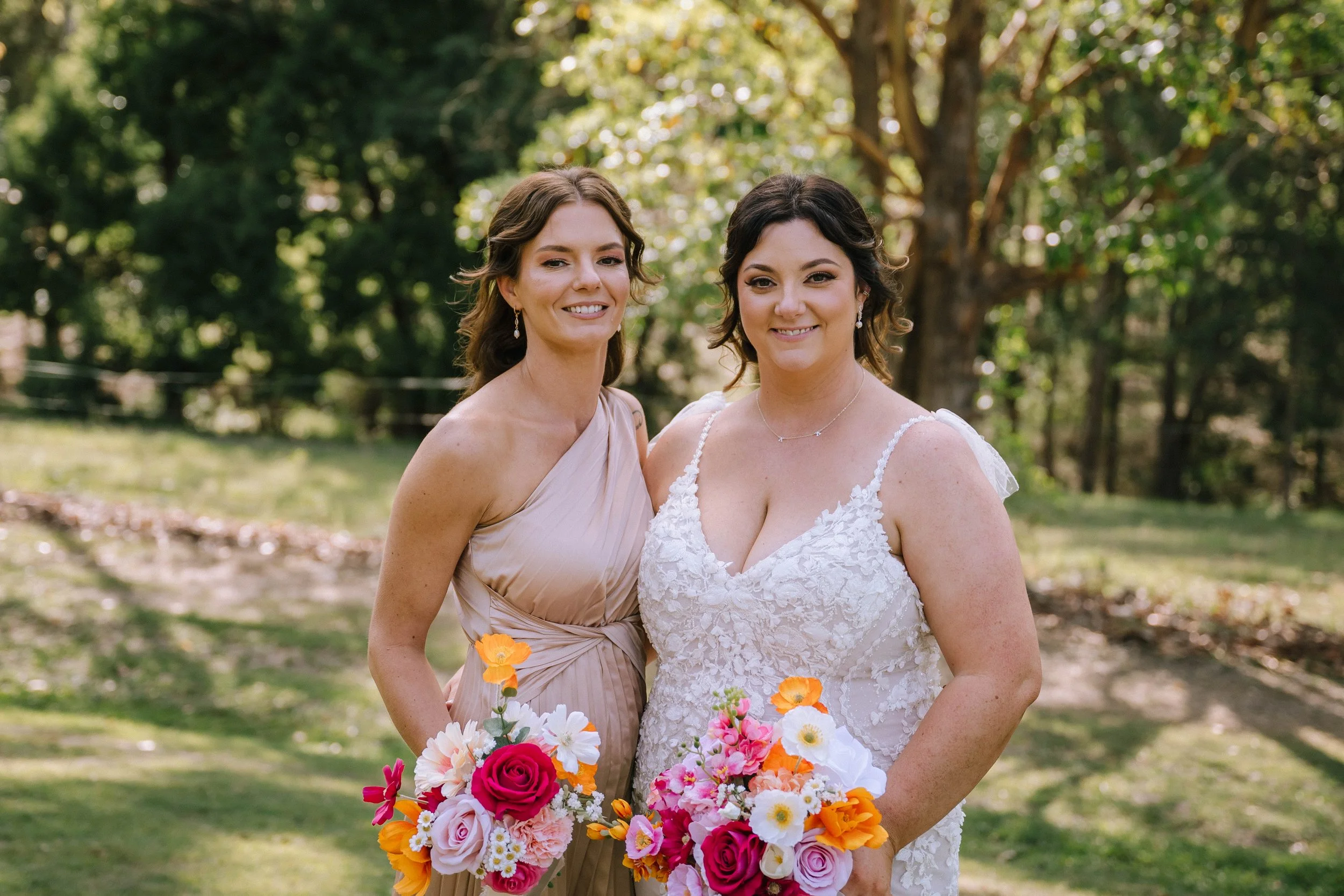 Two women in wedding dresses holding colorful bouquets standing outdoors in a park with trees and sunlight in the background.