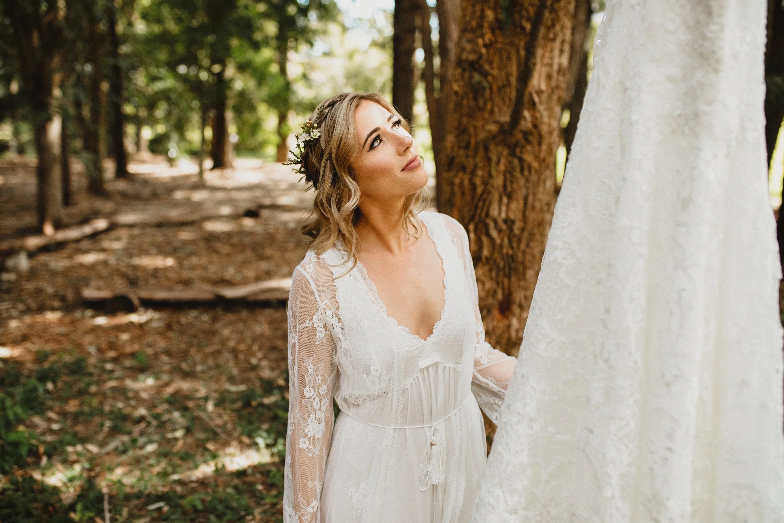 A woman in a white lace wedding dress and floral headband looks up at a hanging wedding dress in a wooded outdoor setting.