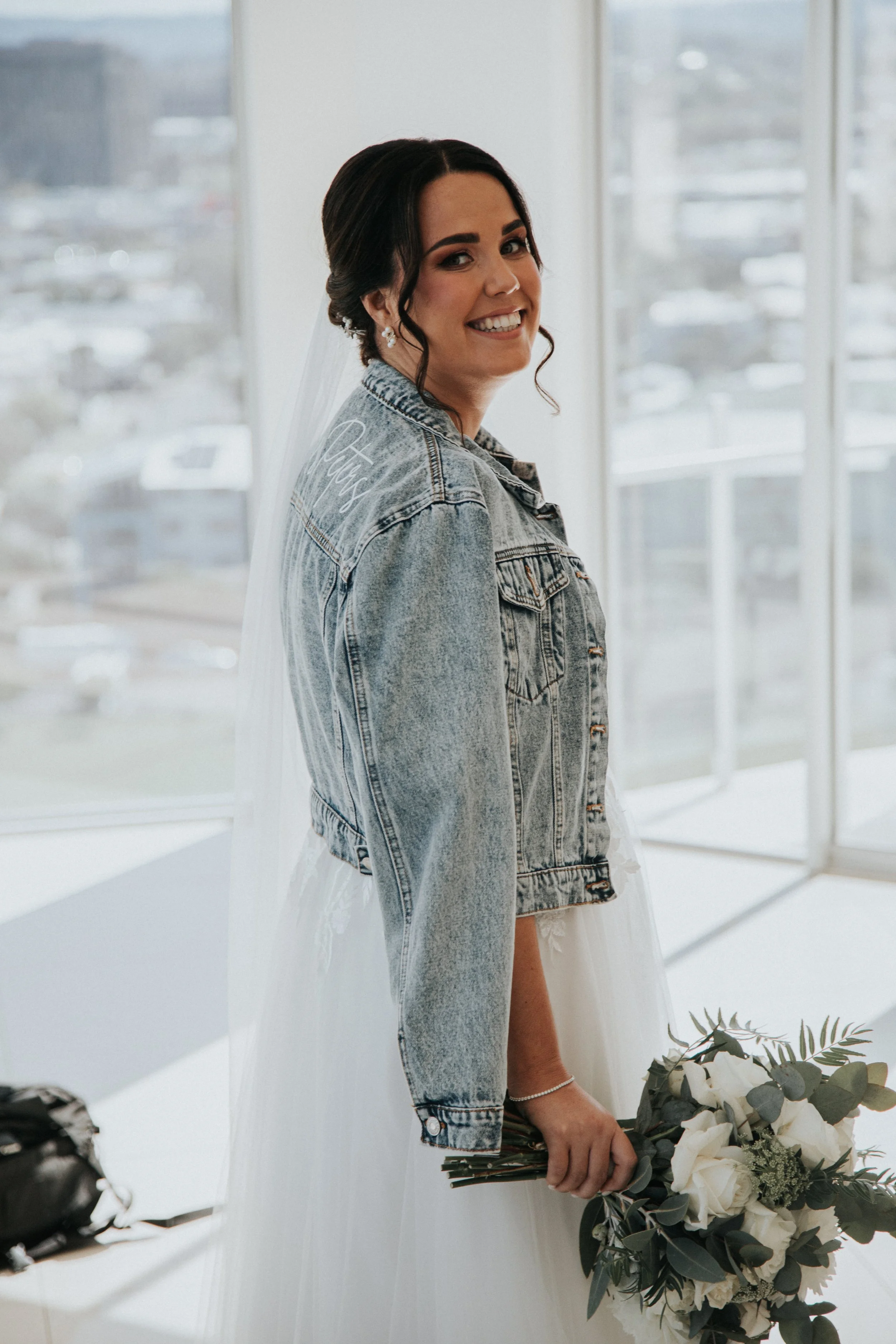 A woman in a wedding dress holding a bouquet of white roses and greenery, wearing a denim jacket, smiling.