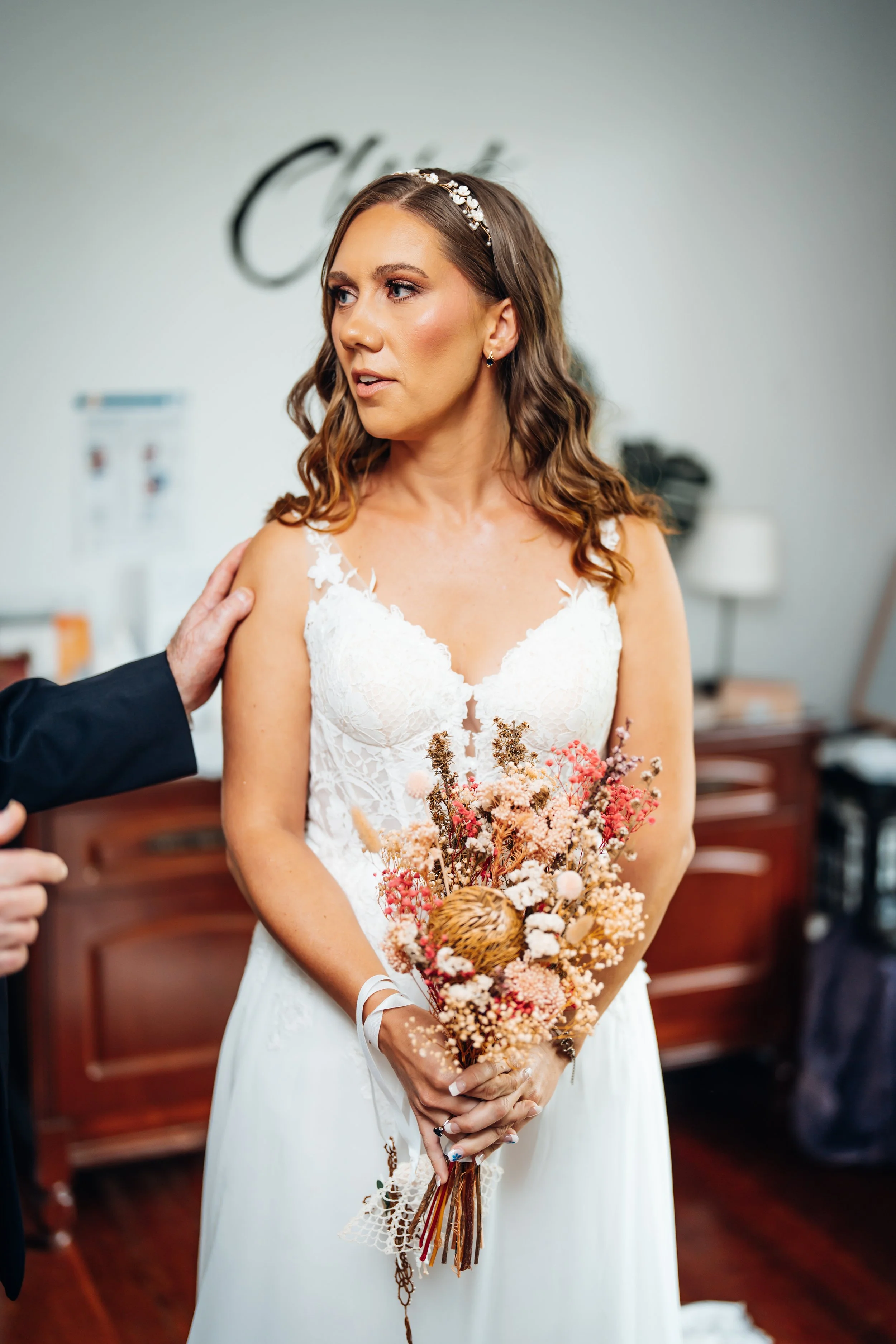 Bridal woman in wedding dress holding bouquet, with a hand on her shoulder, indoors with a blurred background.