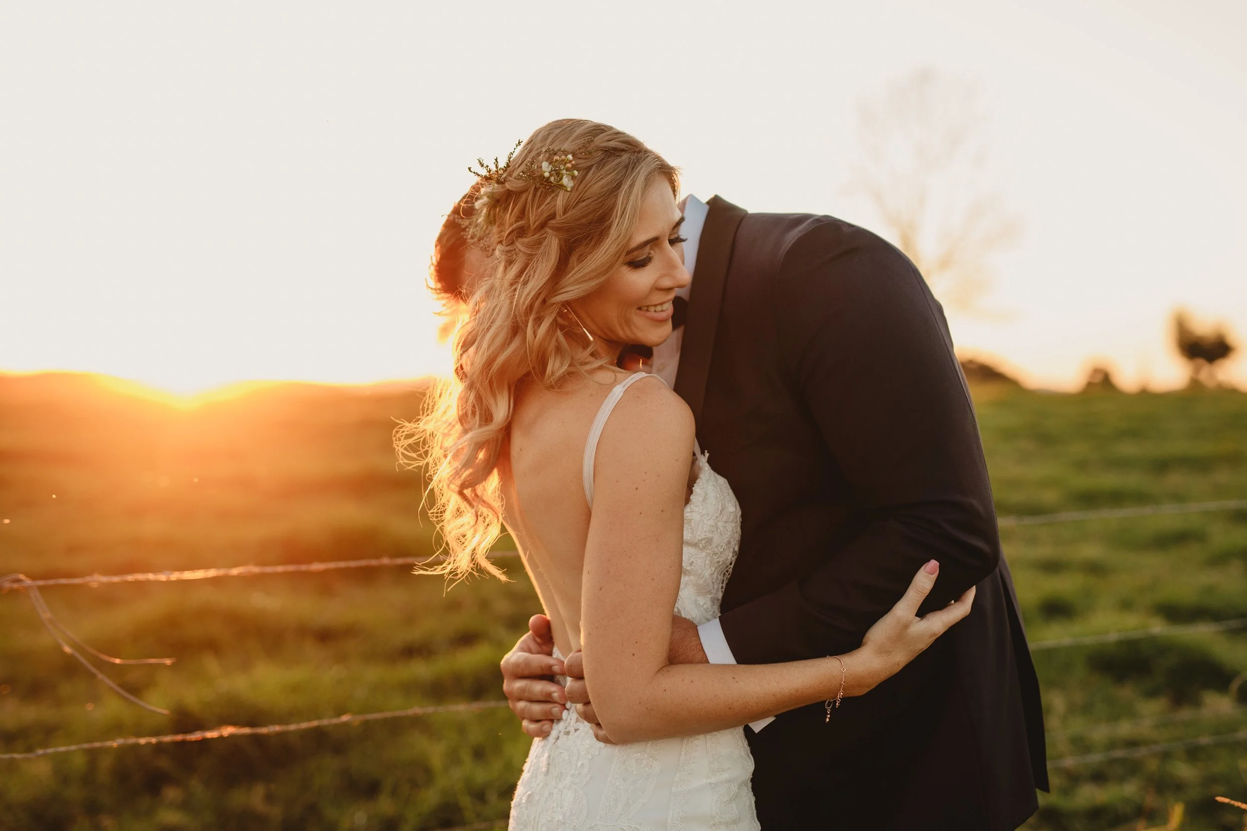 A bride and groom embrace outdoors at sunset, with the bride smiling and the groom leaning into her, in a field with a fence and trees in the background.