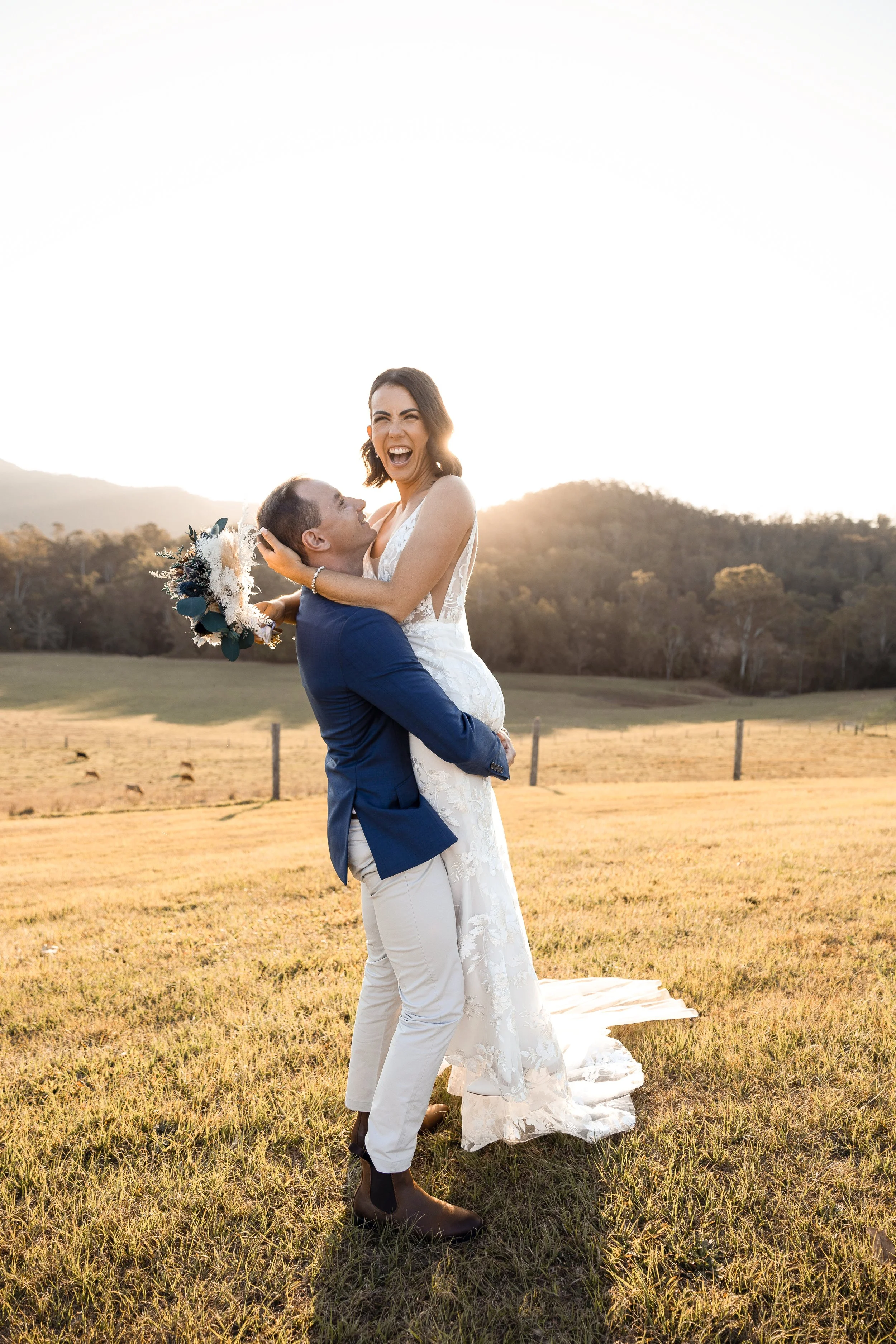 A newly married couple in a grassy field at sunset, with the groom lifting the bride, who is holding a bouquet, both smiling and joyful.