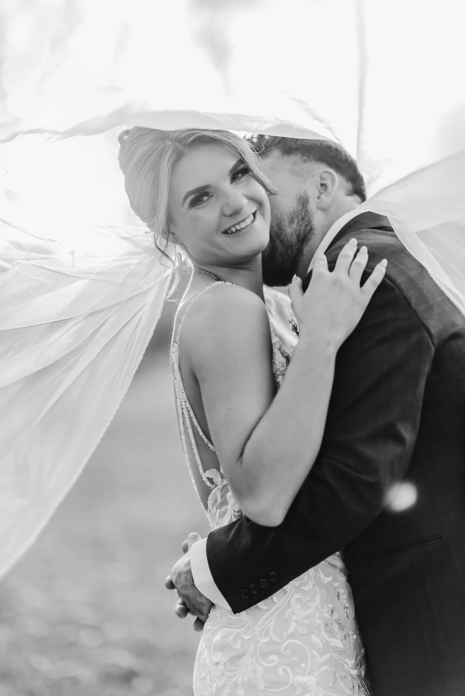 Black and white photo of a bride and groom embracing. The bride has blonde hair and is smiling at the camera; the groom is kissing the bride's cheek, with his eyes closed. The bride's wedding dress has lace details, and the groom is in a dark suit. T