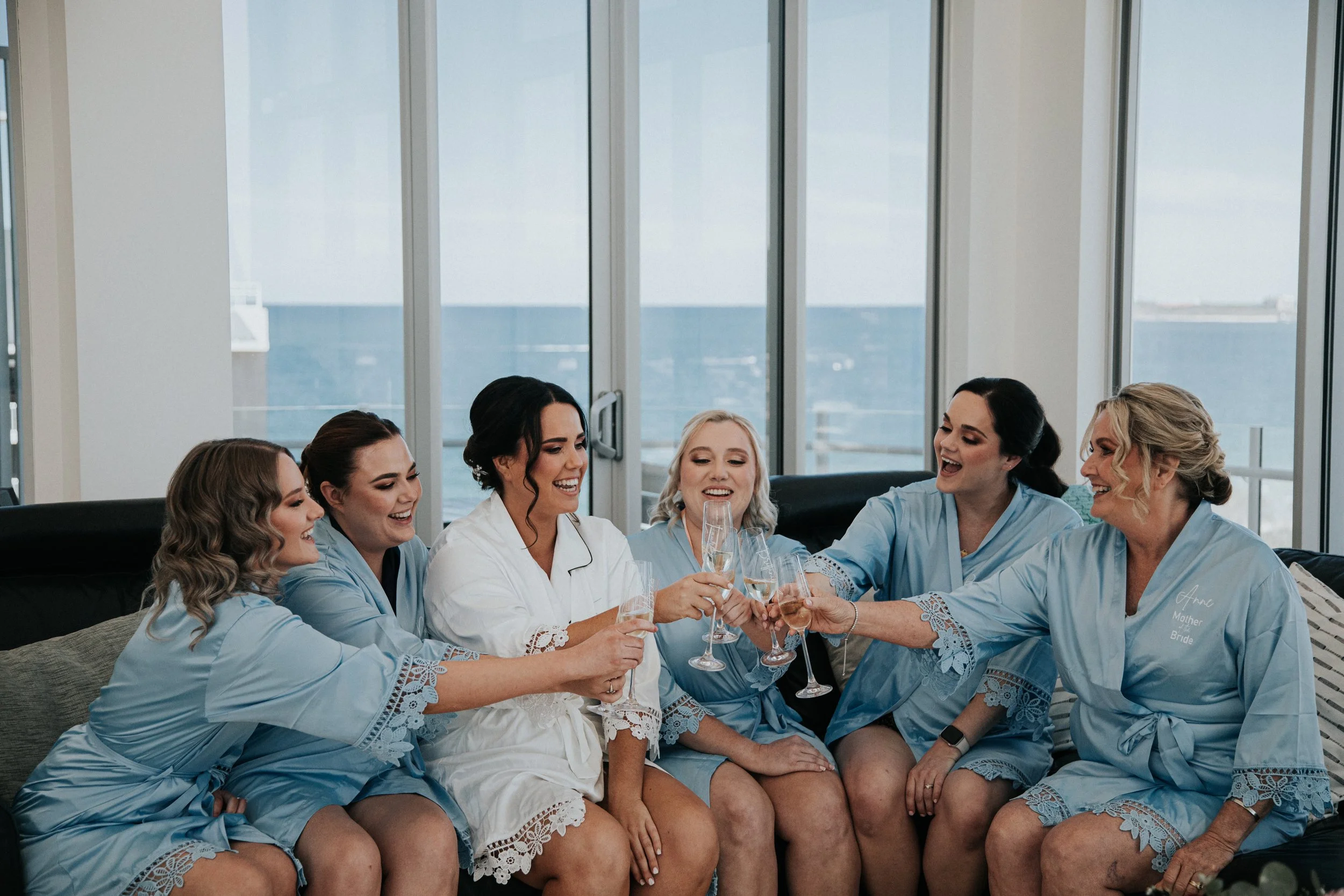 Six women, including the bride in a white robe, sitting on a couch by windows with ocean view, celebrating with champagne glasses.